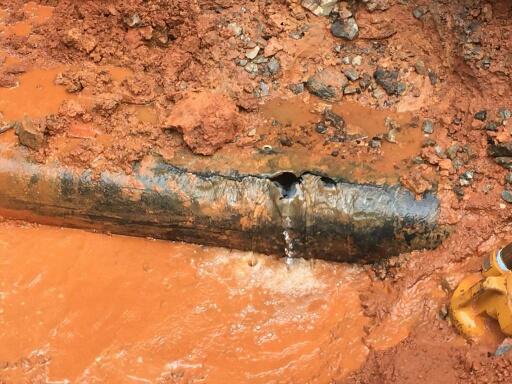 exposed broken cast iron pipe lays broken in mud, a hole leaking water creating a pool of muddy water beneath