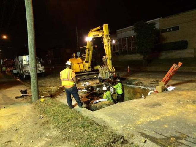 2 utility technicians wearing yellow safety shirts and white hard hats work at night in the dirt of a square pit cut into a road bed as a yellow excavator sits along the edge; nearby stands another technician wearing a yellow safety vest and white hard had