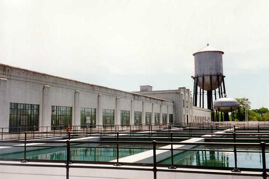 vintage photo of water treatment plants from years ago featuring large 1 story building, several fenced in sediment ponds, and 2 water towers, one tall an cylindrical and the other squat and round