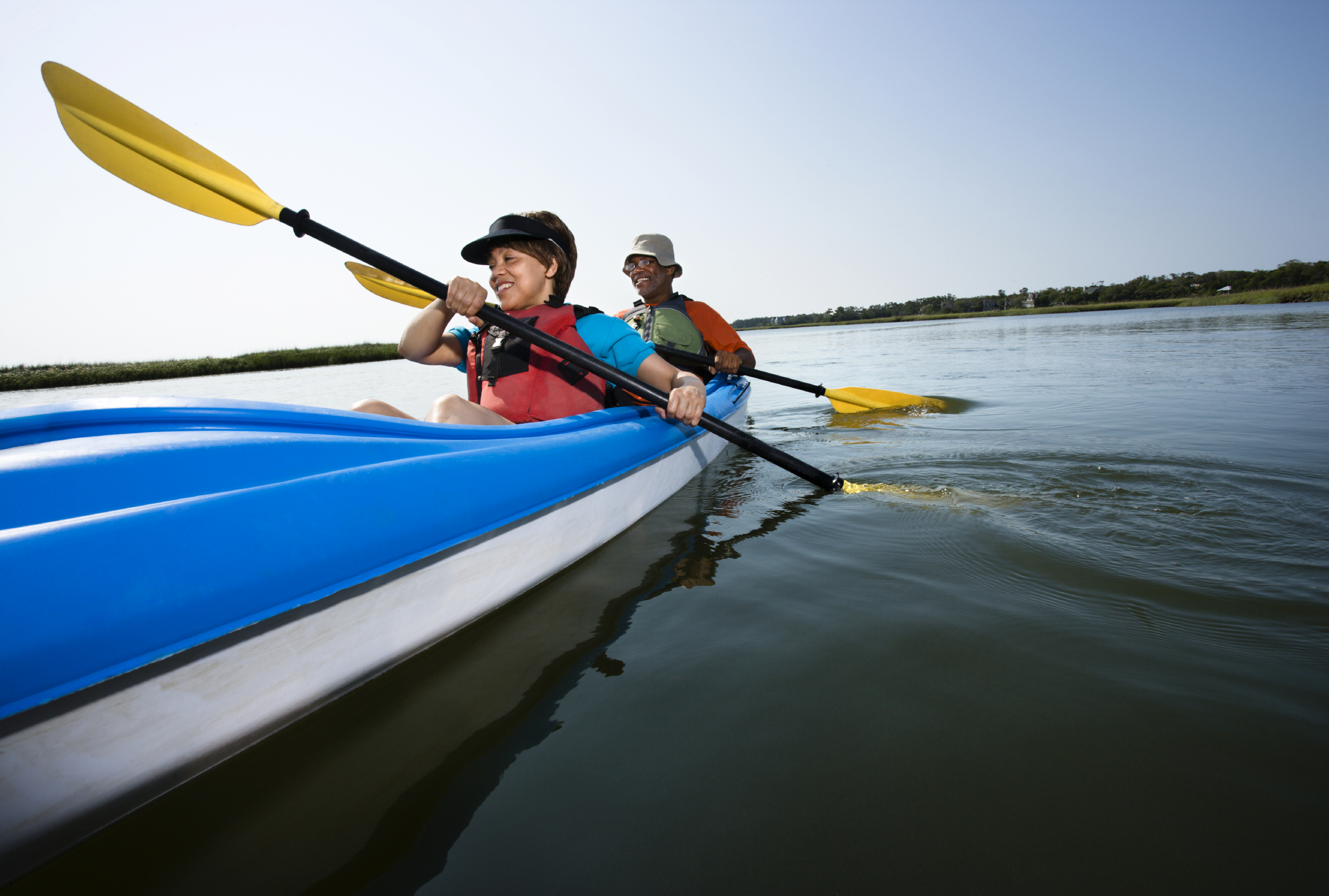 2 people kayak aboard a blue and white kayak using yellow paddles while wearing life jackets making their way across a large pond