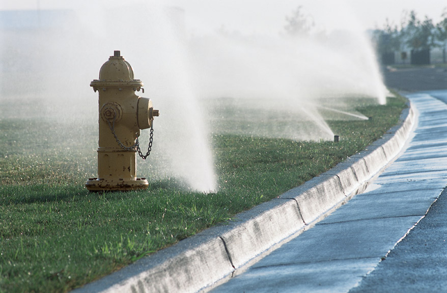 row of black modern sprinkler heads have popped up to spray water on a lawn as a yellow fire hydrants stands in foreground, curbside