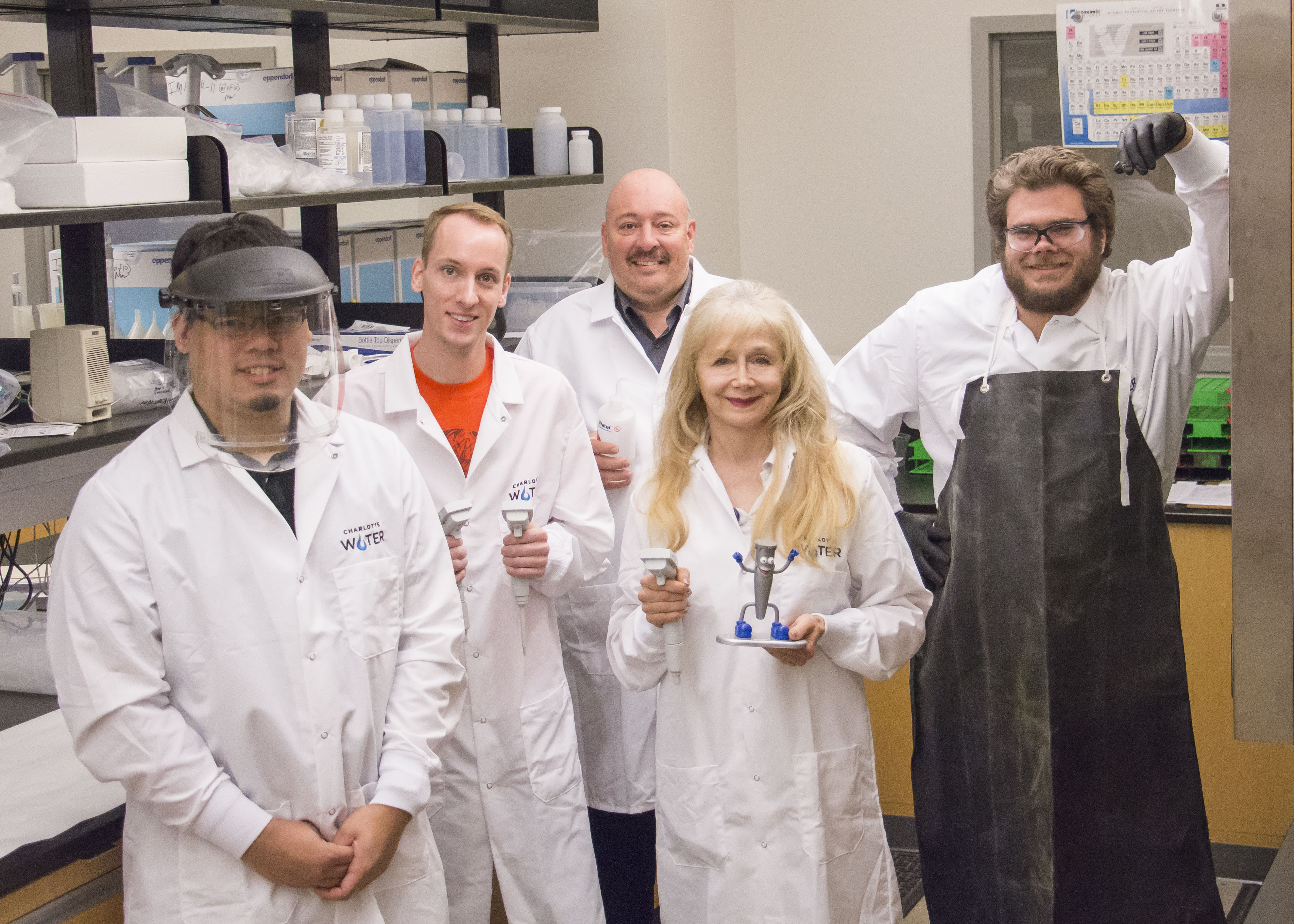 5 lab technicians stand in white lab coats posing for a group photo, one person wears a black rubber apron and another holds a toy doll mascot that is grey with blue boots and gloves