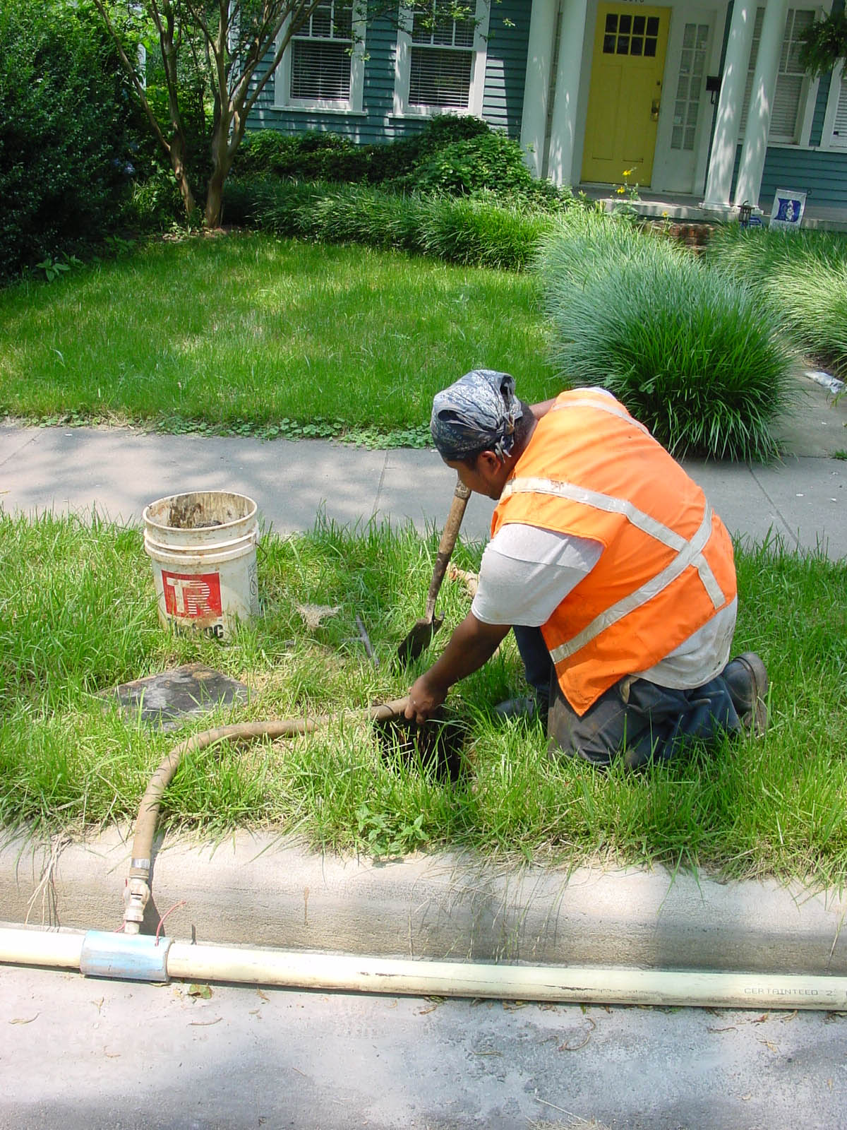 utility technician wearing orange safety vest and blue bandana kneels green grass curbside digging with a trowel to position short section of tan hose to tie into a long white hose running along the nearby gutter