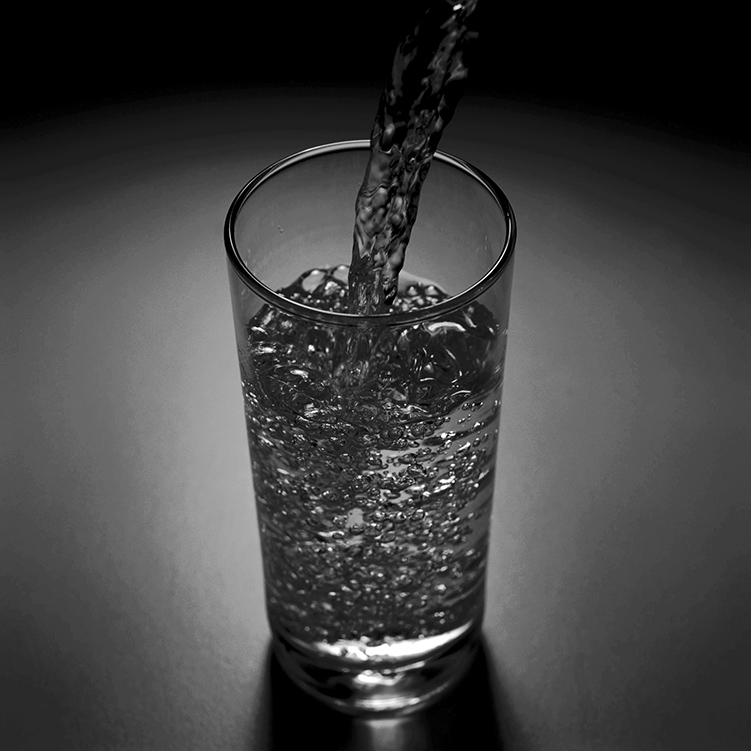 black and white image of a tall glass being filled with water splashing into it