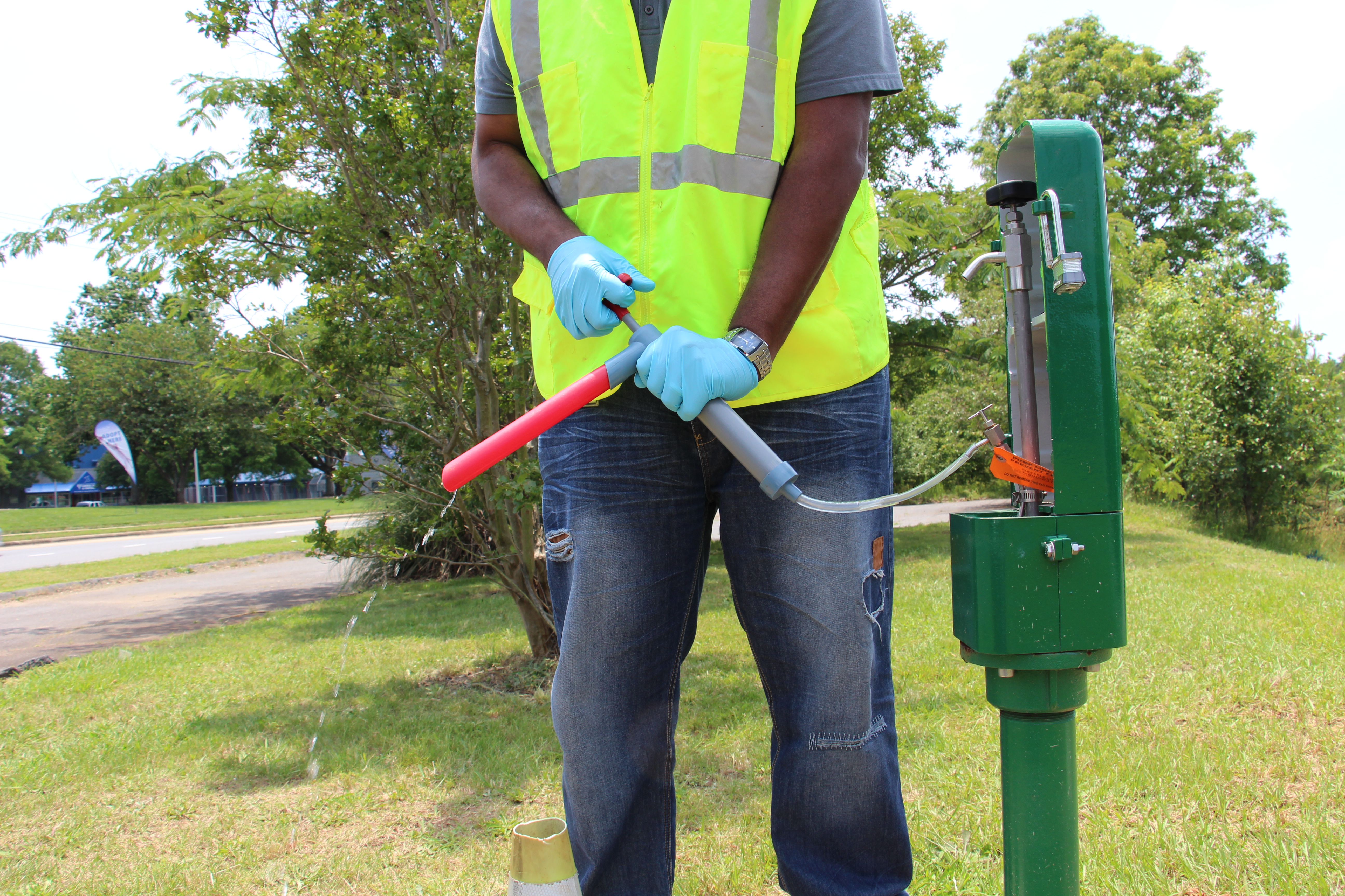 hands of a utility technician wearing blue gloves uses a red and grey hand pump to pump a water out of a green monitoring station into an attached red test tube; the monitoring station is a green rectangular box on a green metal pipe standing in the middle of a grassy area