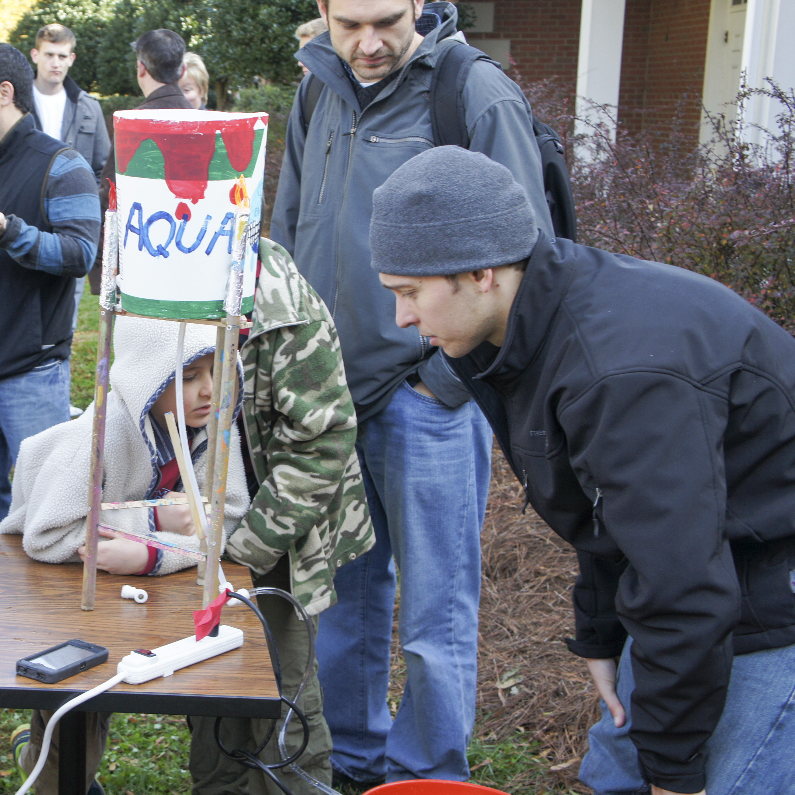 young kid in a white jacket and hood shows off a homemade model of a water tower on a a table as adults review