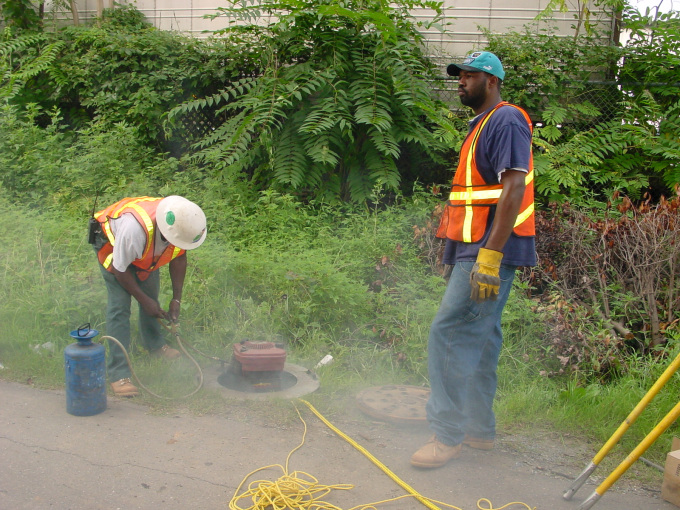 2 utility technicians wearing orange safety vests, one with a white hard hat and other with teal baseball hat spray around manhole access point with a blue spray tank