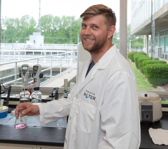 lab technician wearing white lab coat poses for photo while holding small beaker with a light pink liquid in it as he stands near a picture window featuring the water treatment plant sediment ponds