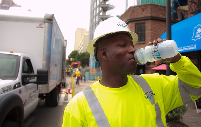 utility technician wearing white hard hat, yellow safety shirt cools off on a city street with a sip of water from his Charlotte Water branded water bottle