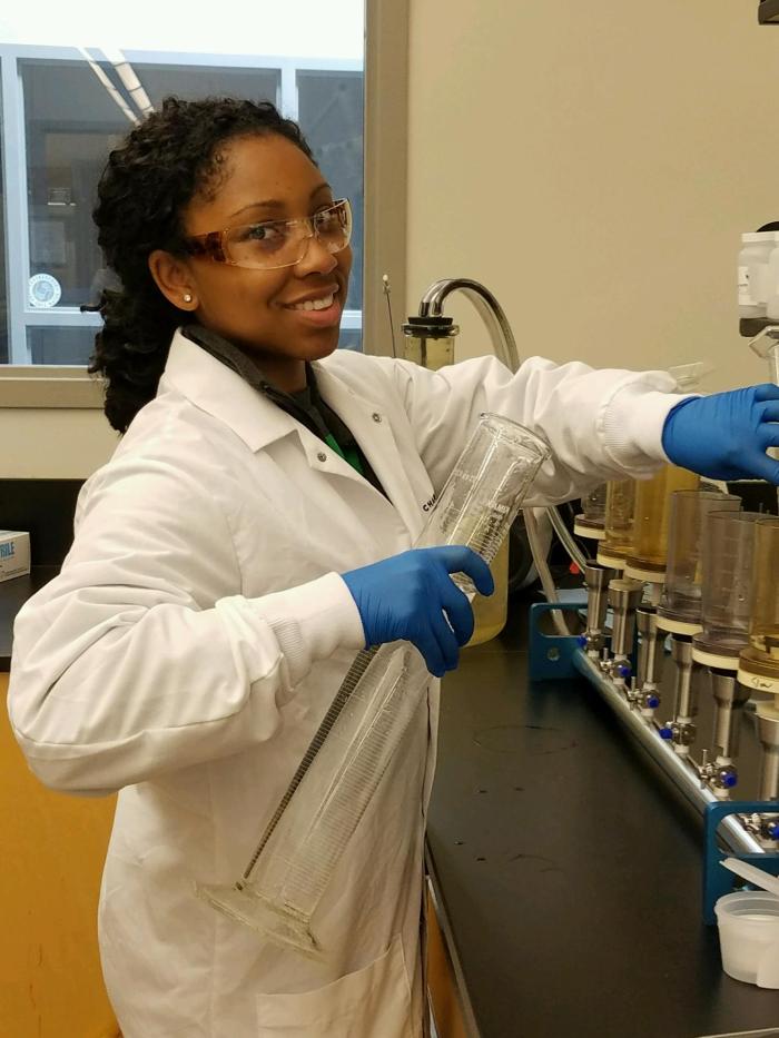 lab technician wearing white lab coat, blue gloves and safety glasses holds a very tall, cylindrical measured beaker as she stands next to a row of glass and metal sample stations