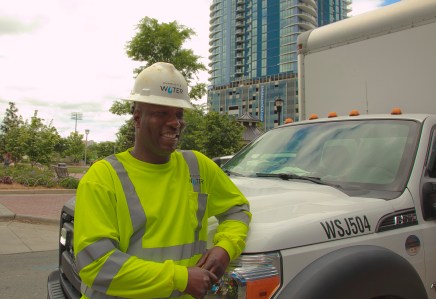 utility technician wearing yellow safety shirt and white hard hat leans against white utility box truck with blue and glass apartment tower behind him