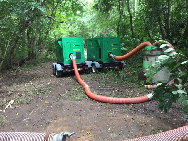 2 portable green pump generators pump water using orange hoses from off camera and into a tall concrete sewer access point