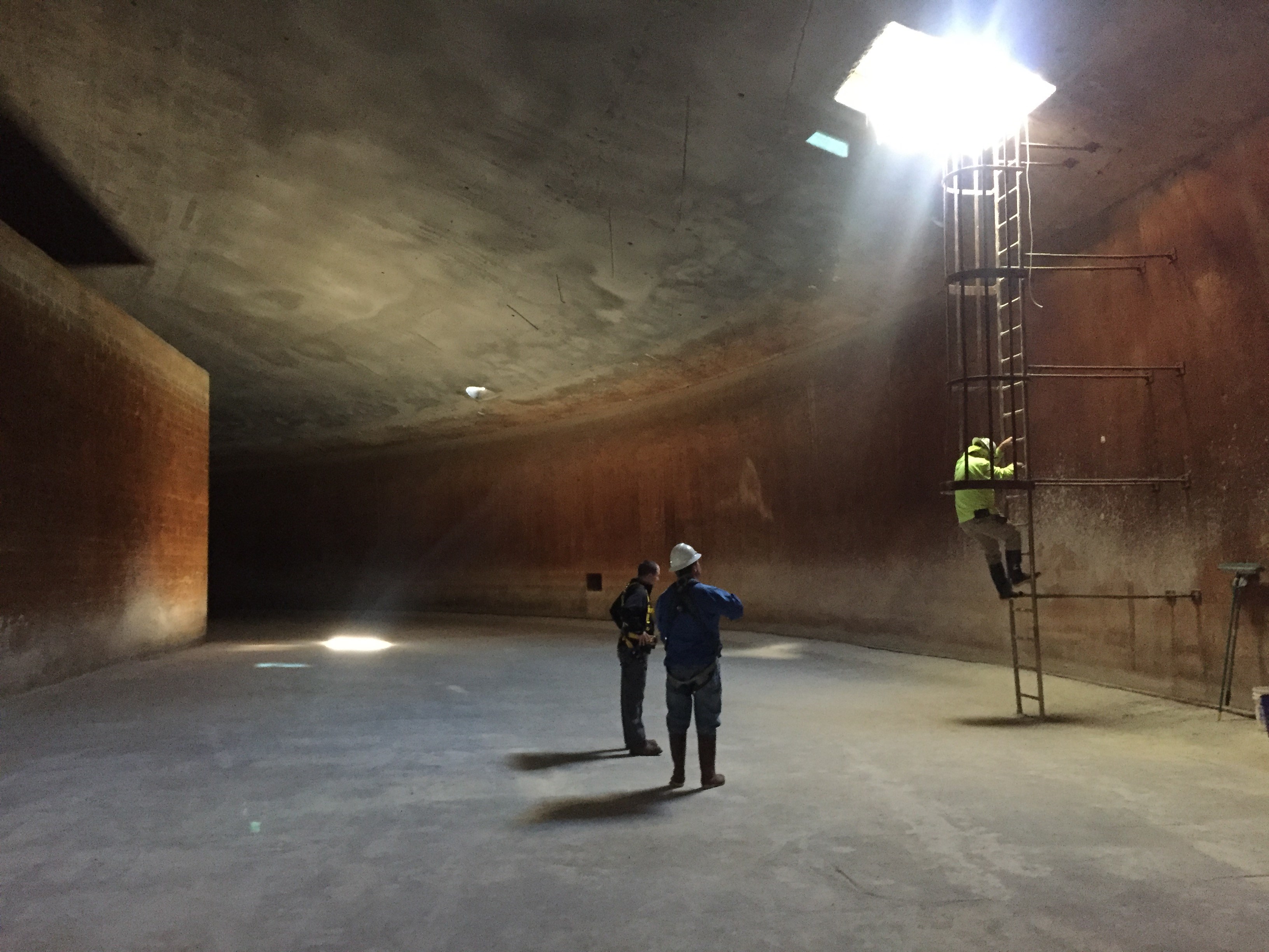 3 utility technicians inspect the inside of an enormous water treatment tank by climbing down inside as sunlight from the hatch above illuminates the brown and concrete cavernous space