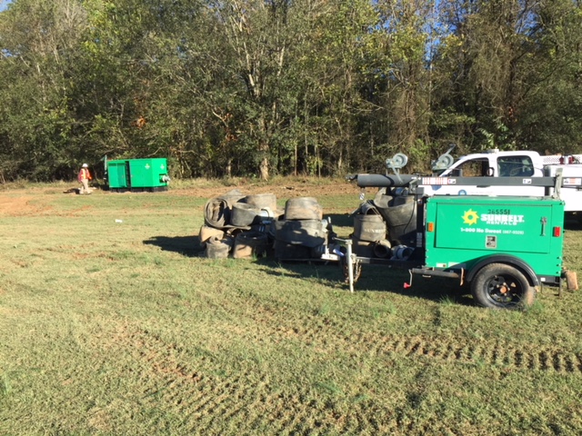 portable green generator is parked on soggy field of green grass next to a stack of black cylinders, a white utility pick up truck; a large green metal container sits along tree line in distance