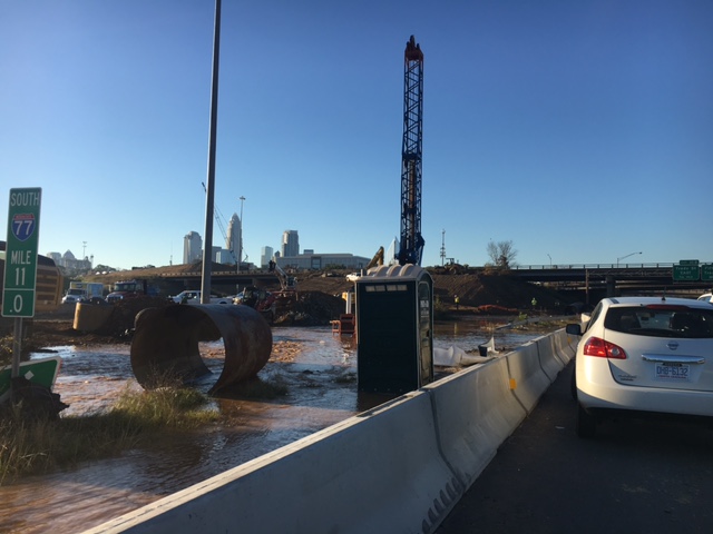 flooded job site along a interstate 77 with single section of metal pipe, black porta-potty, black crane and a view of the city skyline in the distance
