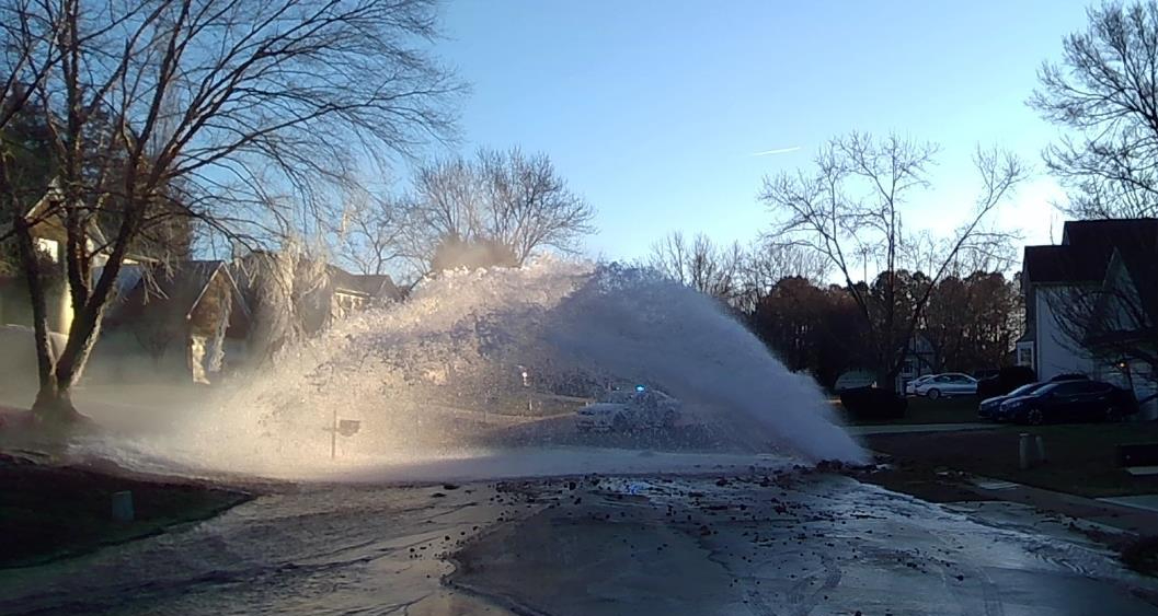 geyser of water plumes up from a curb along a suburban street flooding the roadway with water and mud