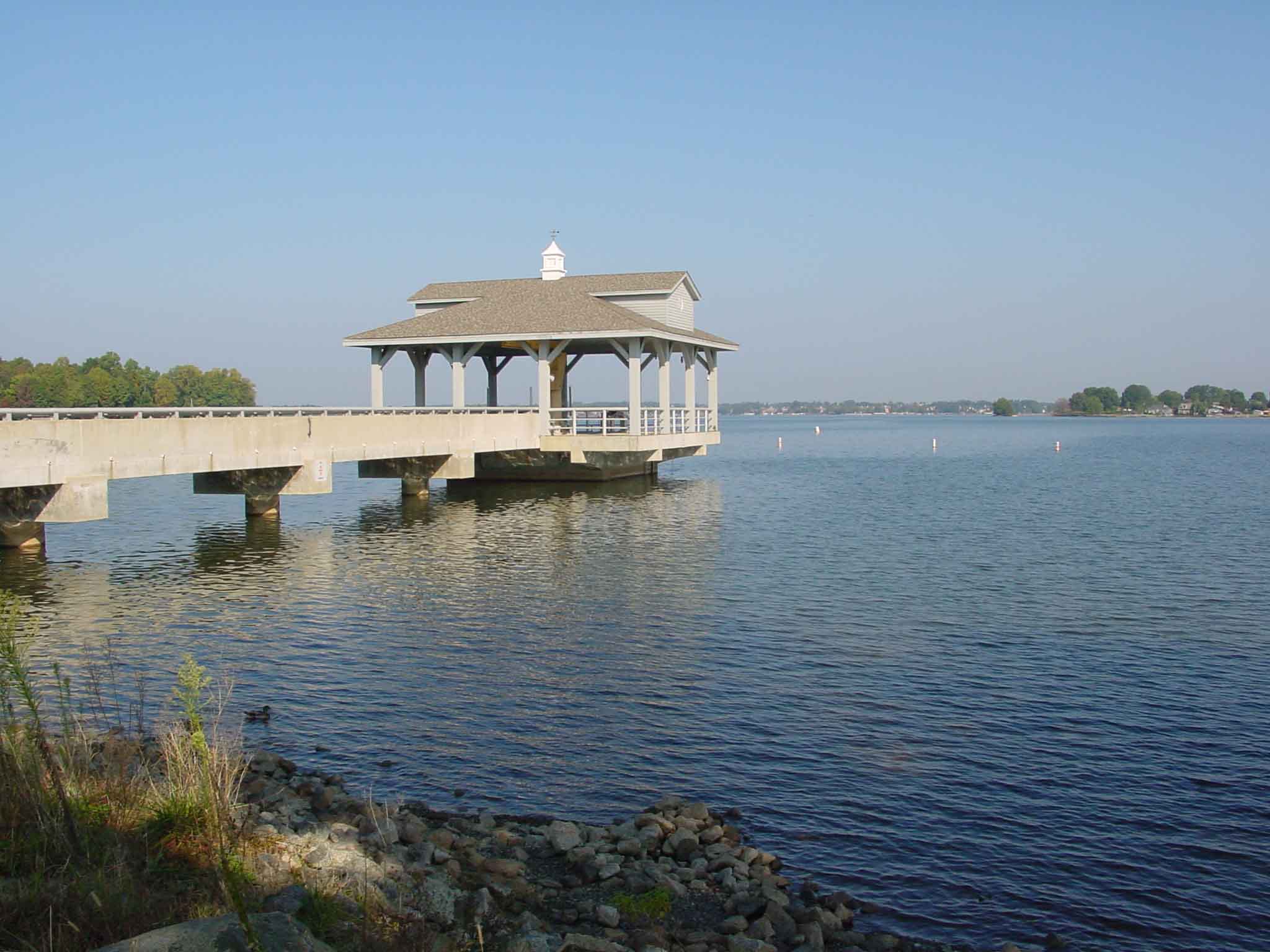 white and tan gazebo sits at the end of a concrete pier with a panoramic view of a harbor with blue waters and shoreline behind it