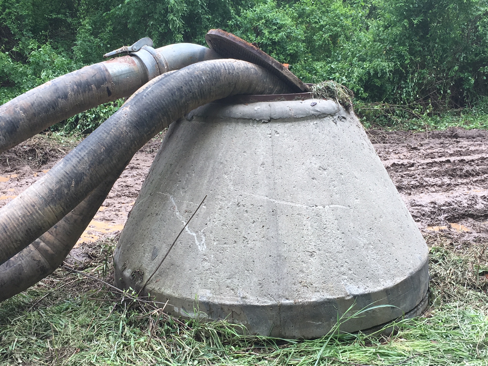 3 enormous black and muddy hoses have been stuck into the opening of a tall concrete sewer access point via an open cast iron manhole cover