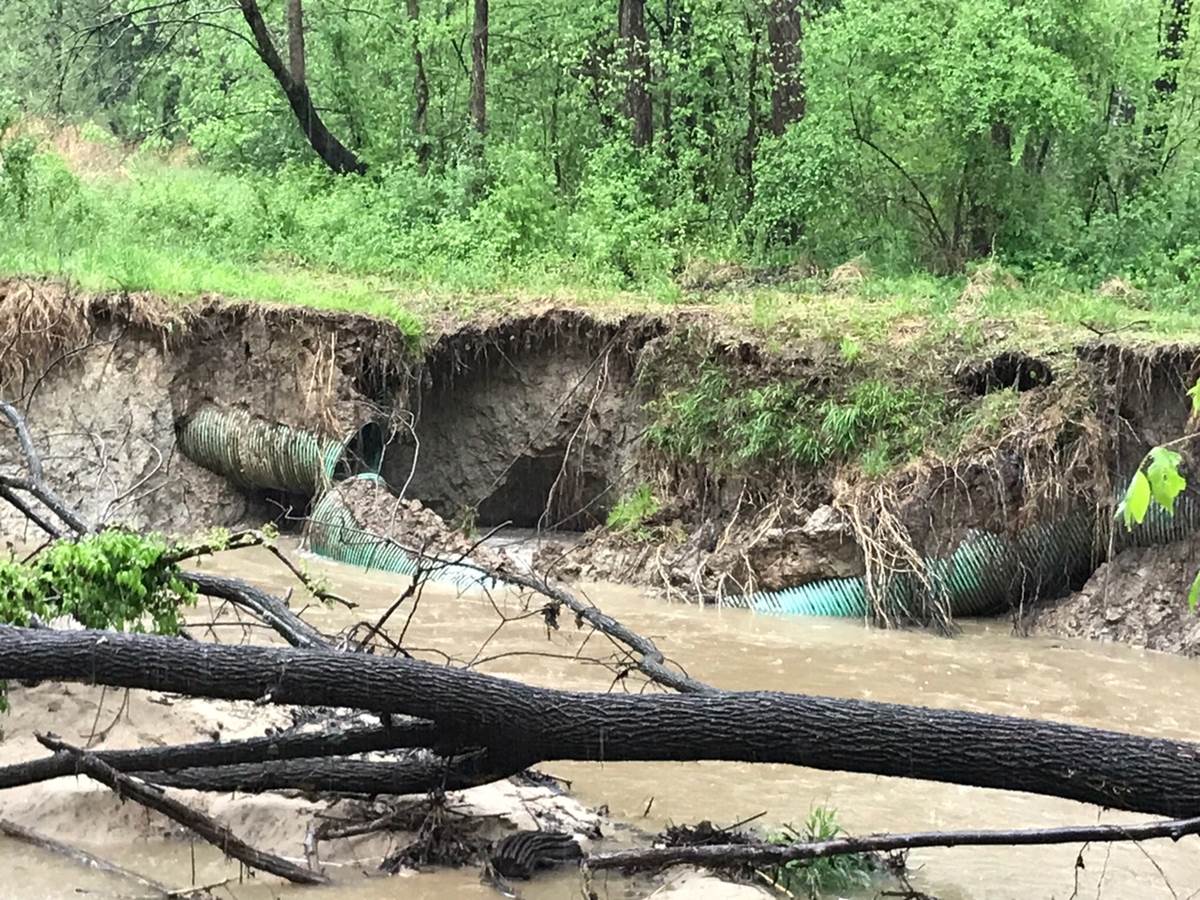 section of large green flexible pipeline is broken due to a flooding event in the adjacent stream; mud, tree debris are strewn about