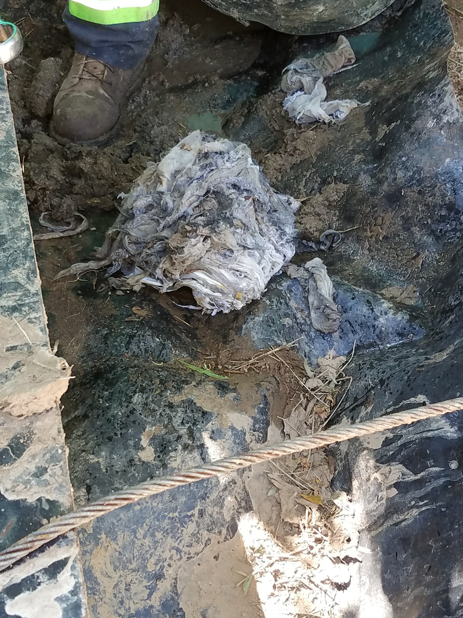 view of plastic trash, debris and mud in a sewer access basin, booted feet of a technician stand near by