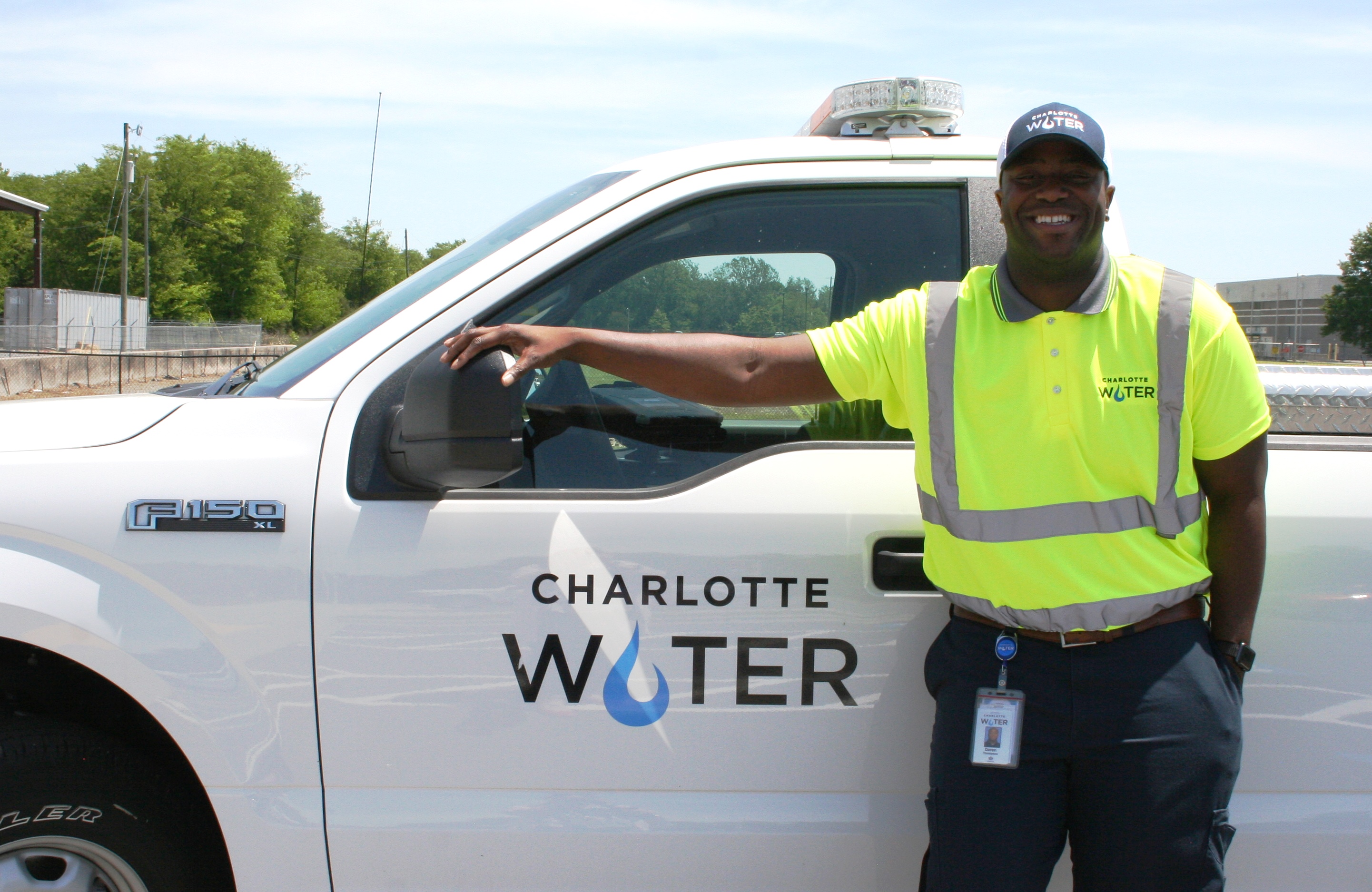 utility technician wearing yellow polo safety shirt, blue baseball cap both with Charlotte Water logos, stand smiling for photo next to white utility pick up truck also with Charlotte Water logo