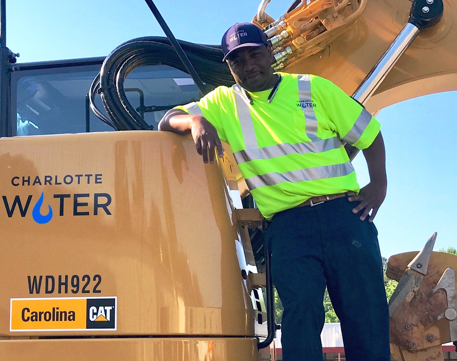 utility technician wearing yellow safety t-shirt and blue baseball cap both with Charlotte Water logos smiles as he leans against a yellow CAT excavator with Charlotte Water logo