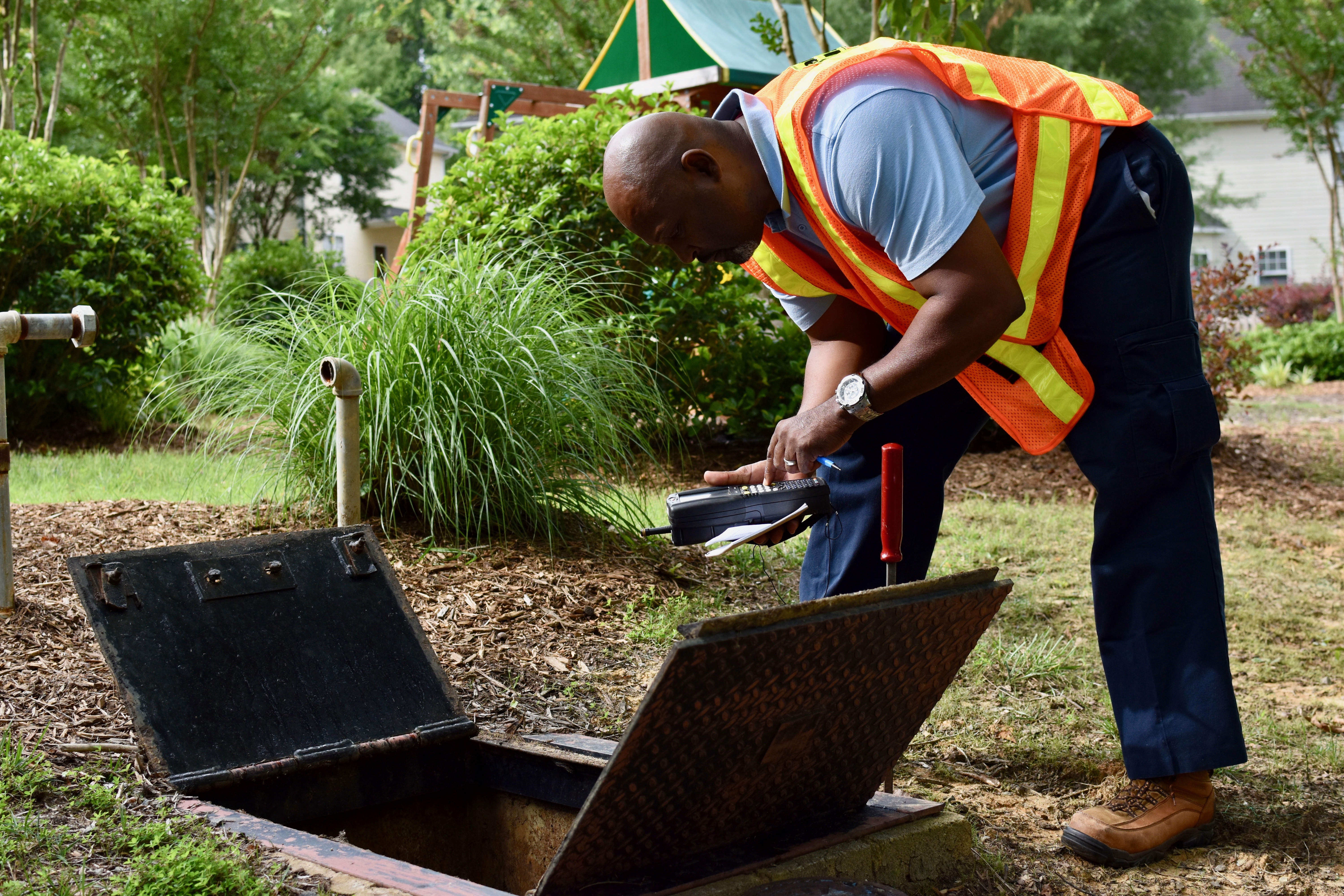 adjacent to a residential backyard, a utility technician bends down over open metal doors of an in-ground service port, as he reviews what is inside he punches information onto the keypad of his black hand-held meter reading device; his red handled tool key sticks up nearby