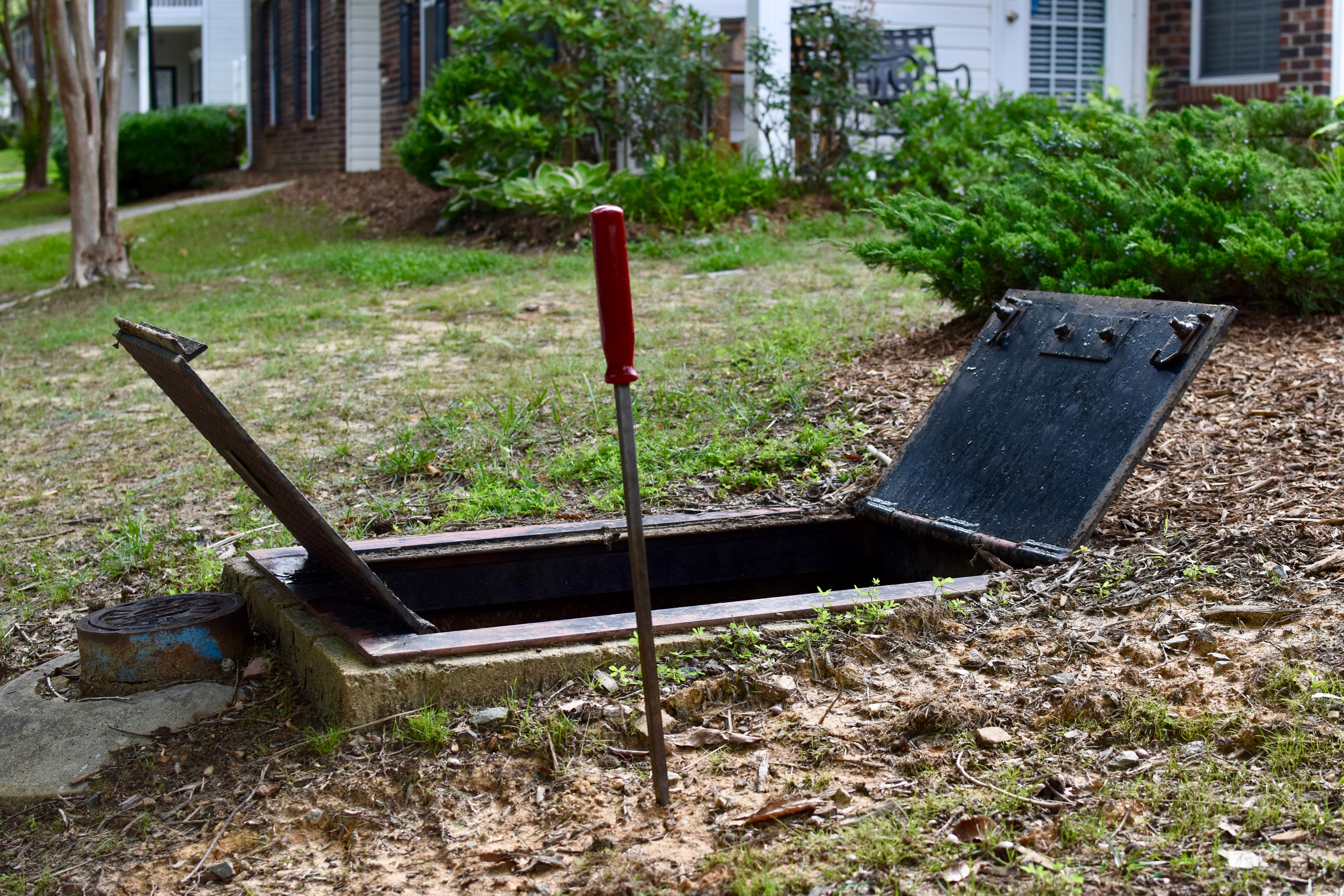 near a residential home, view of 2 open metal service doors reveal an open rectangular port in the ground, nearby is a utility company's red handled tool key and a small, blue cast iron water department access point