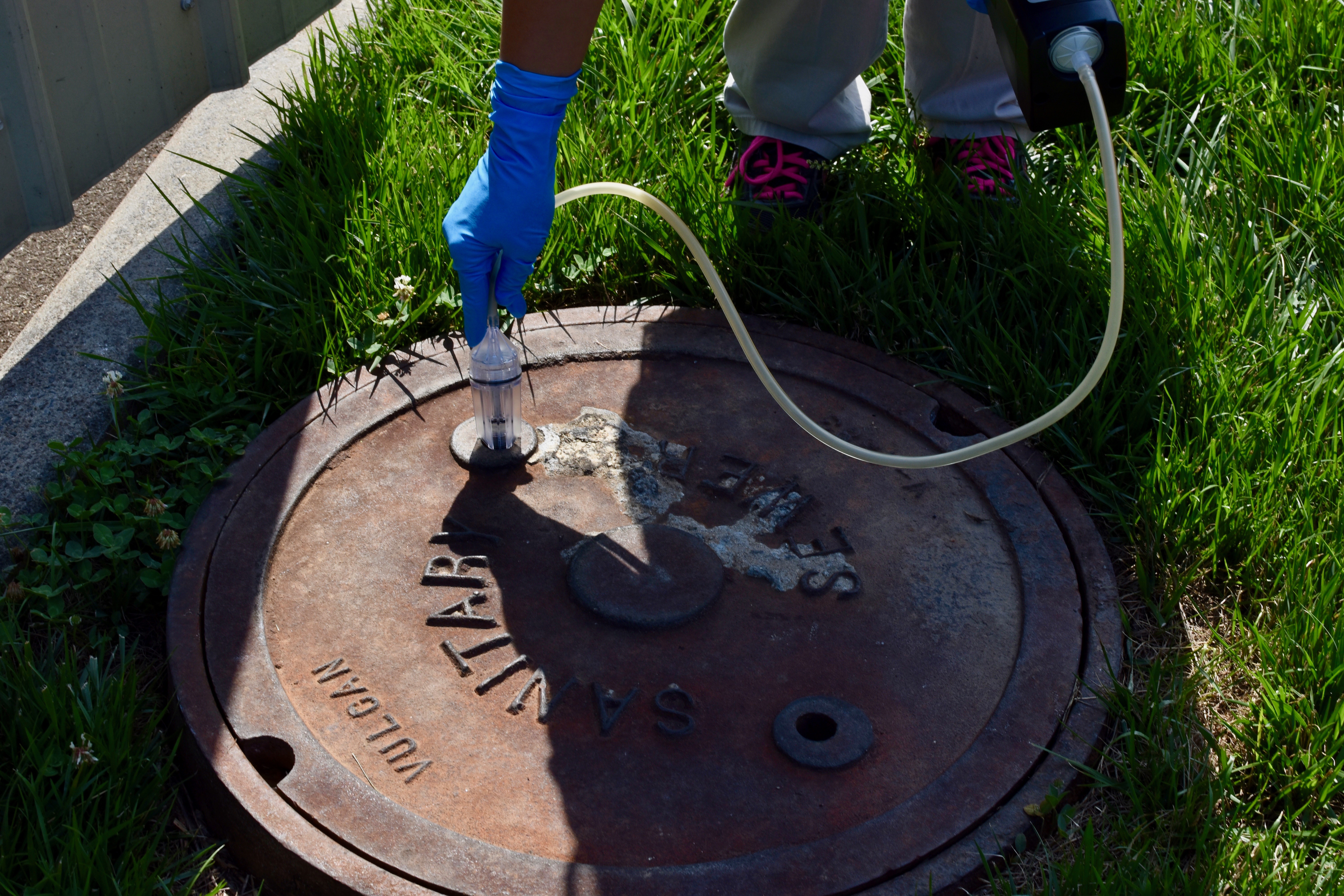 blue gloved hands insert a clear tube into a service port of a brown cast iron manhole cover, the other hand holds the receiving part of a small hose connected to the tube, the lid of the manhole cover says Sanitary Sewer