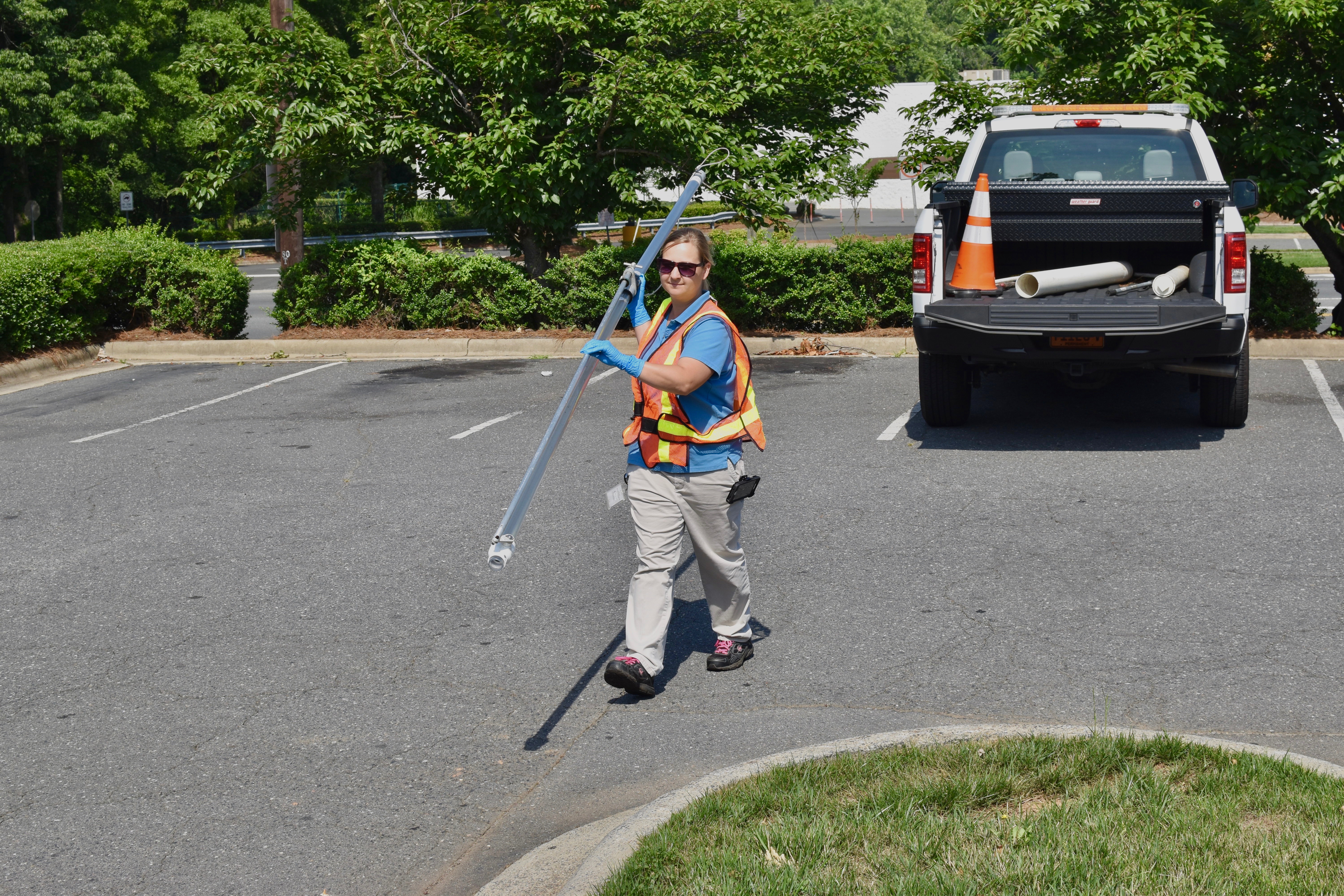 utility engineer wearing orange safety vest walks from open bed of white pick up truck carrying a long tube across parking lot