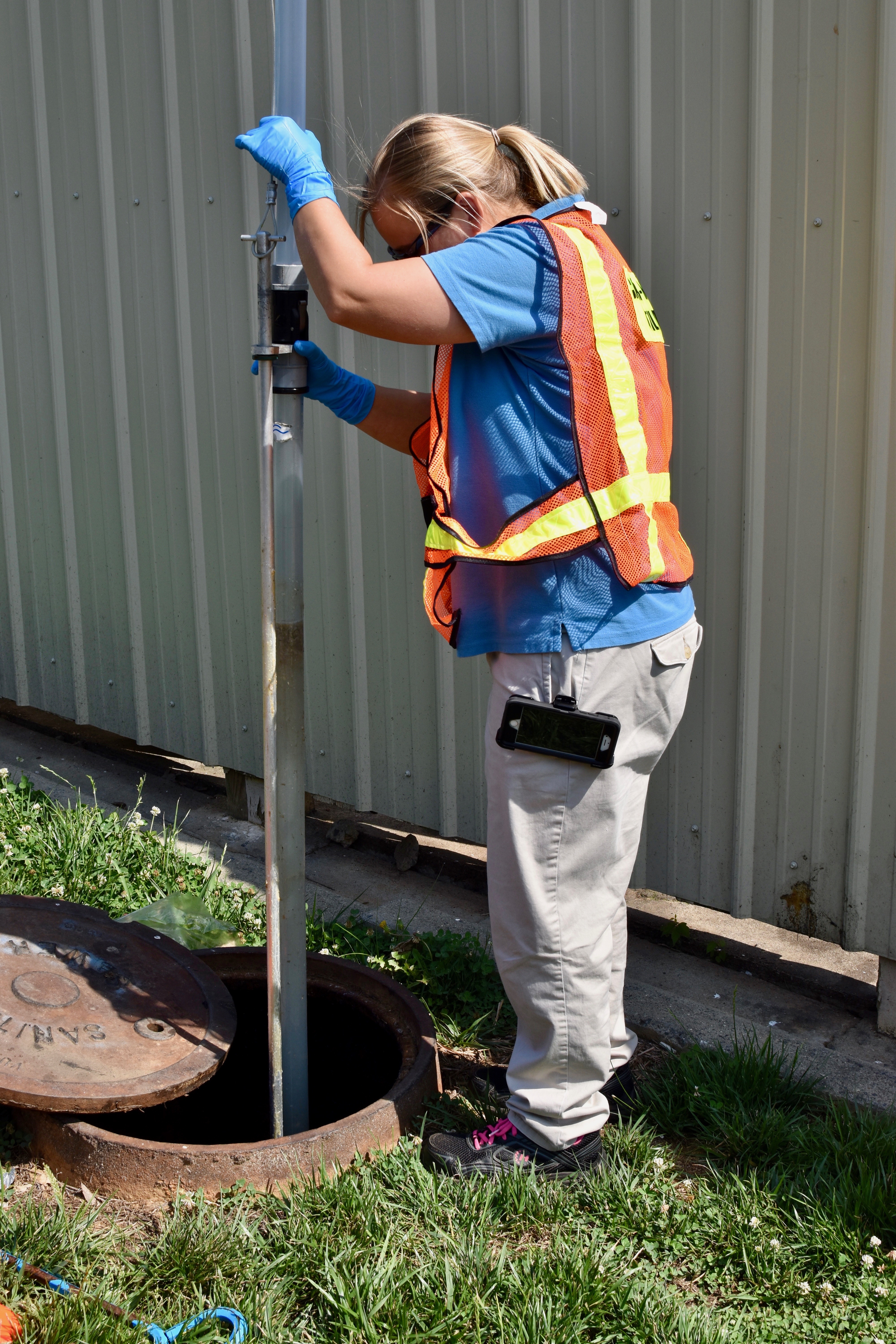 utility engineer with blonde ponytail stands looking down cast iron opening of a manhole while she inserts a clear tube tool to extract liquid samples from below