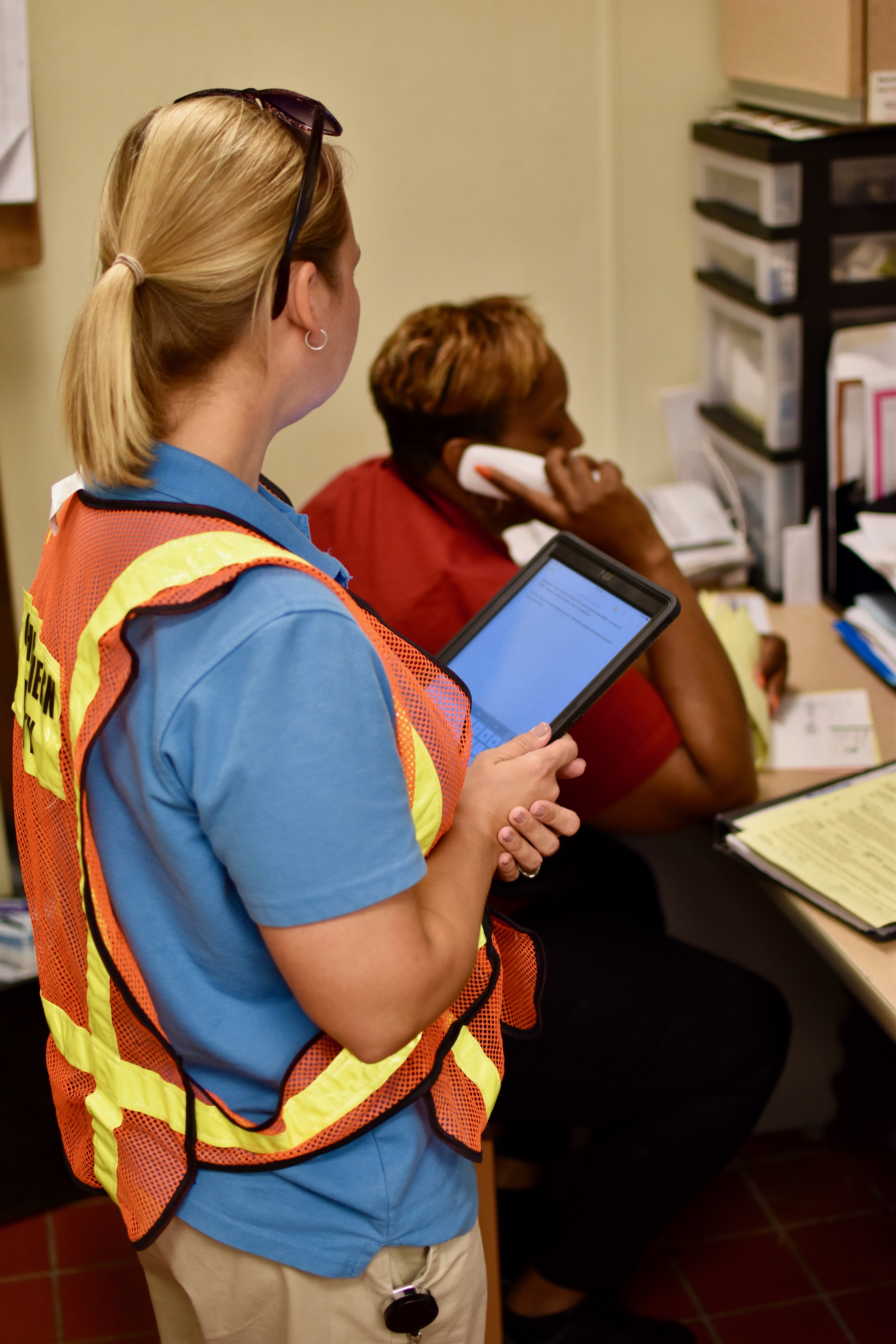 utility engineer with blonde pony tail wearing orange safety vest holding black ipad stands and waits behind a co-worker seated at a desk on the phone