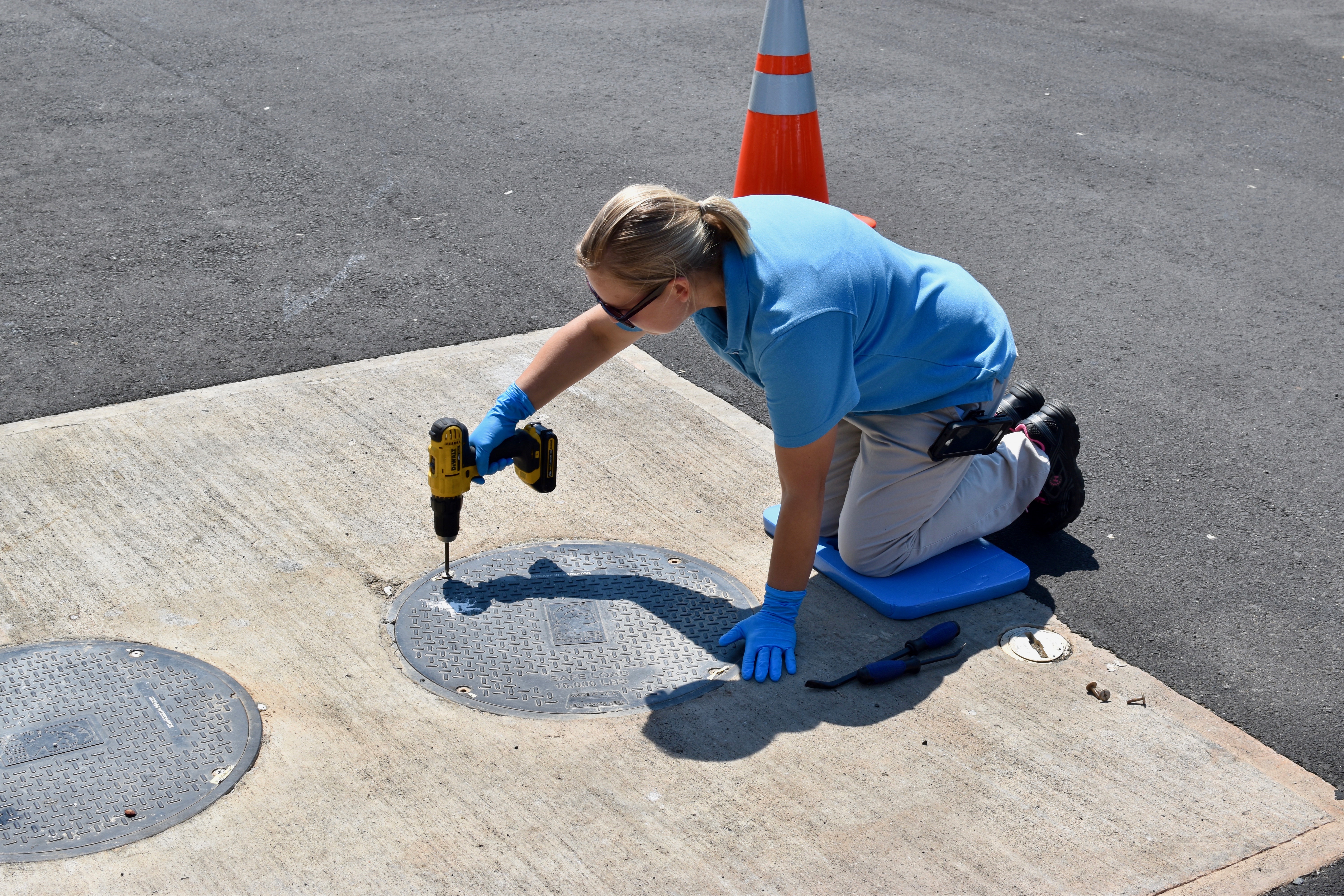 orange traffic cone nearby and yellow power screwdriver in hand, utility engineer with blonde ponytail kneels on blue cushion as she unscrews the lid of a manhole cover