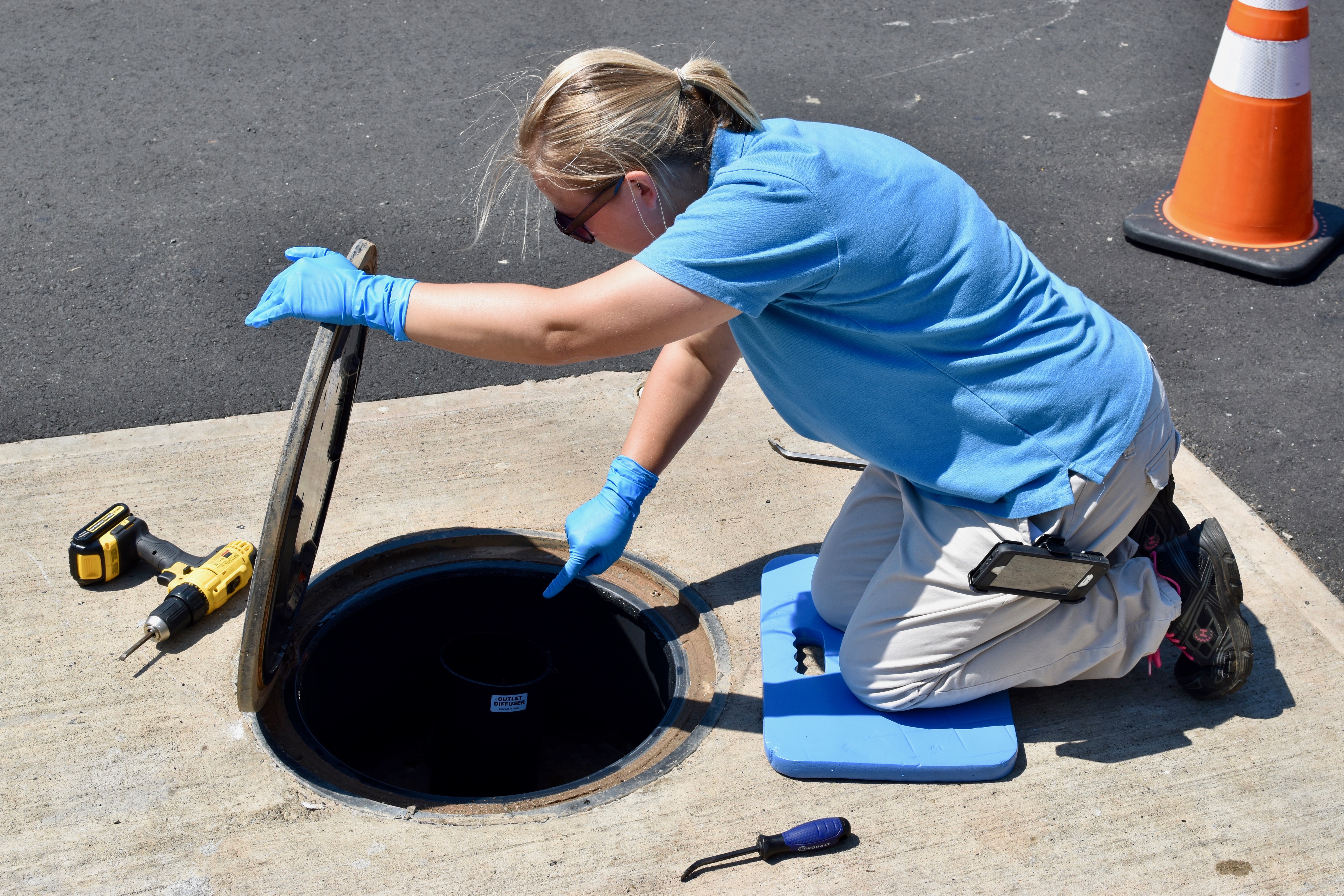 orange traffic cone and yellow power screwdriver nearby, utility engineer with blonde ponytail kneels on blue cushion as she lift up the lid of a manhole cover and looks down inside