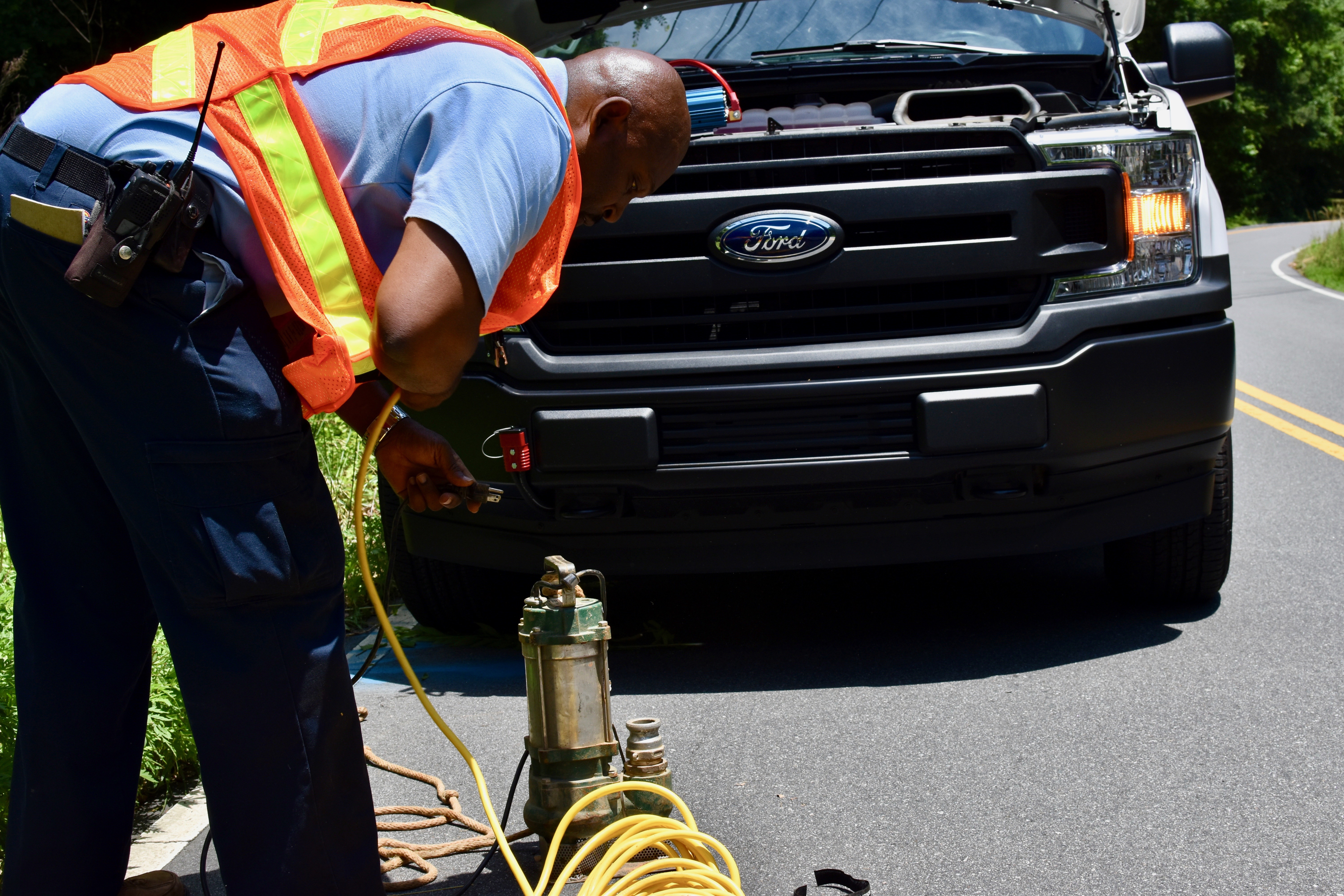utility technician wearing a yellow safety vest leans down on a street in front of a parked truck and sets up a portable green sump pump with yellow power cord coiled nearby