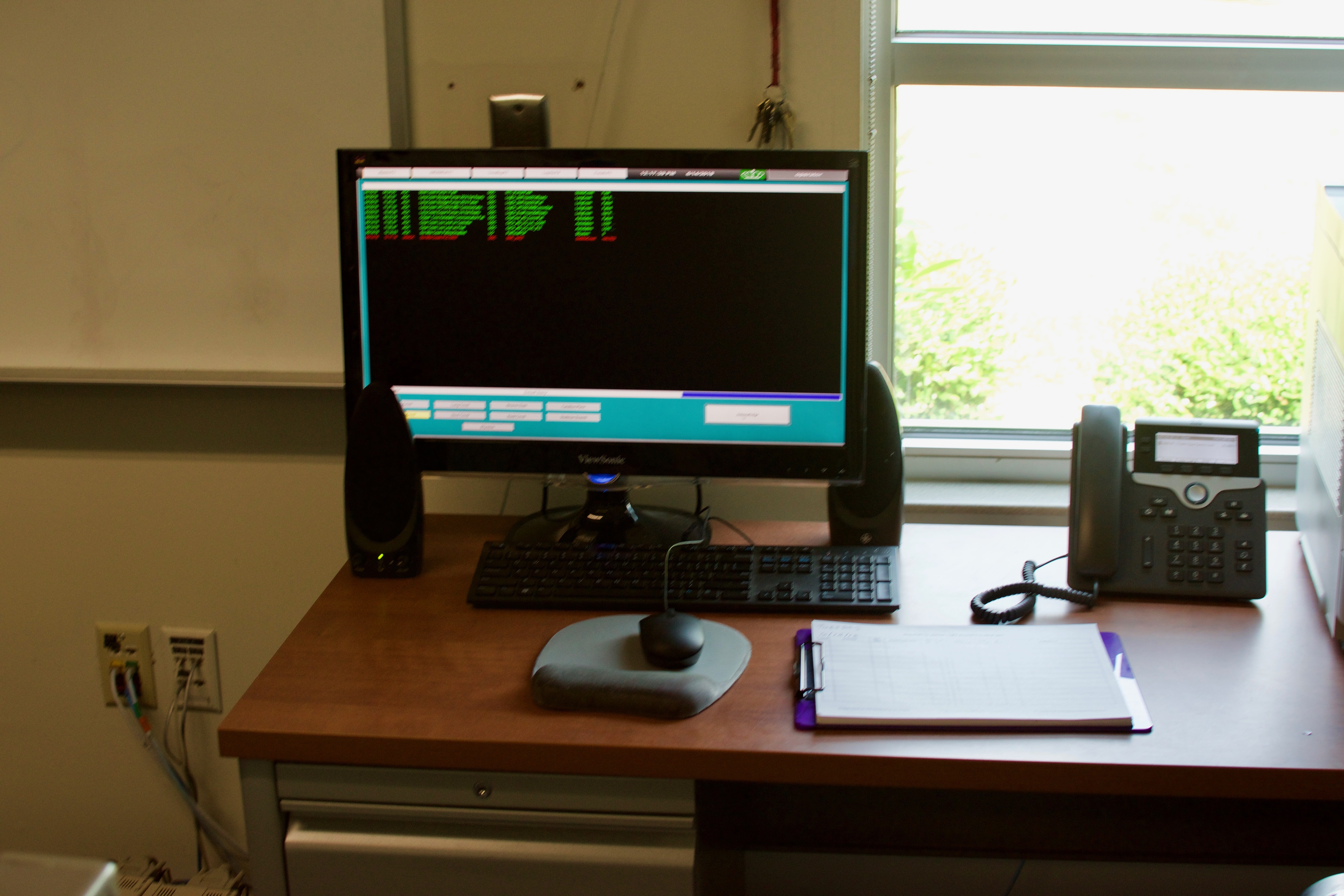 computer monitor sits on a wood desk with a desk phone, mouse, mousepad and purple clipboard, it displays a list of data in green font