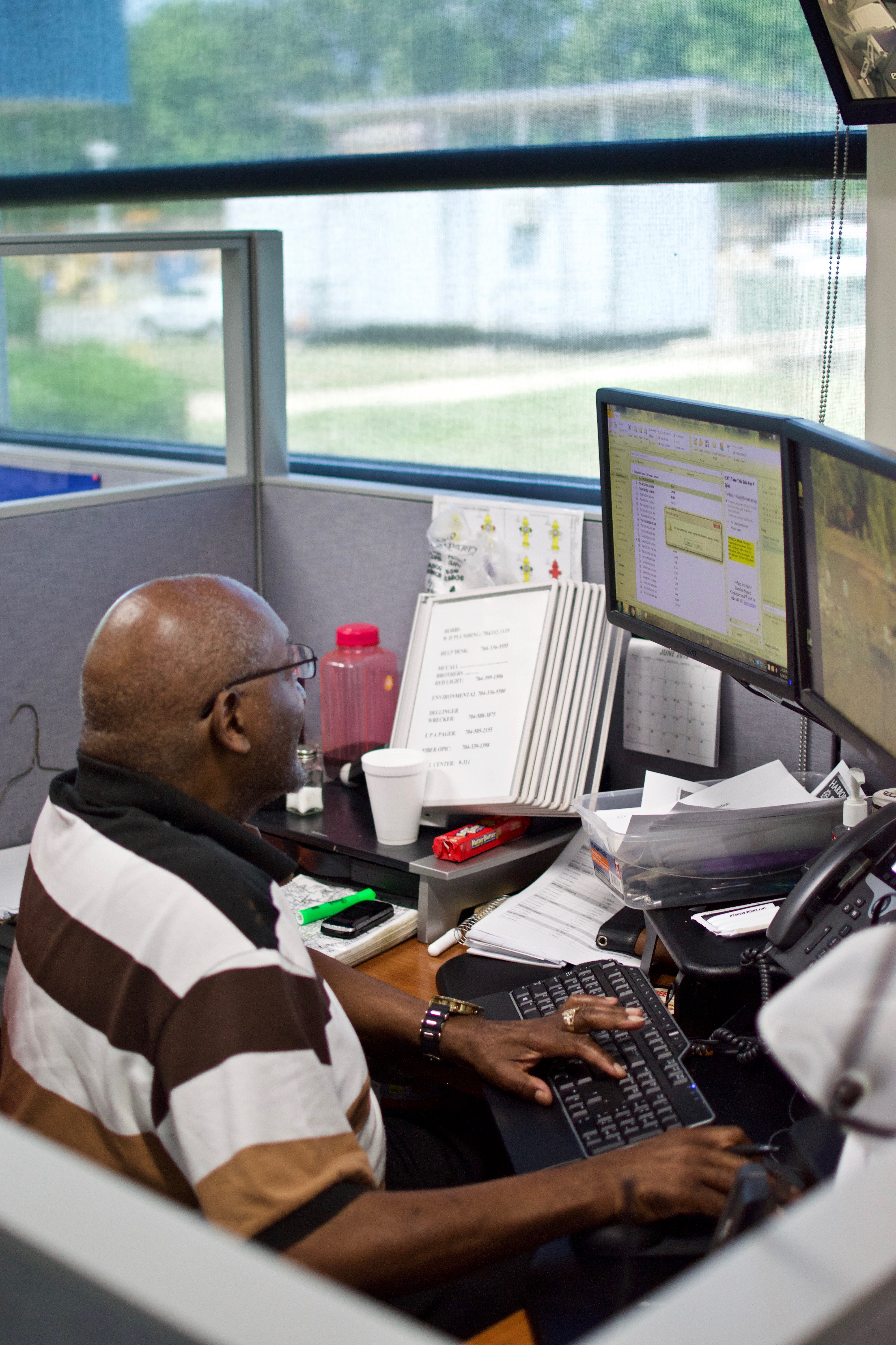 utility employee sits in a grey cubicle with windows, he types on black keyboard while reviewing data on two monitors