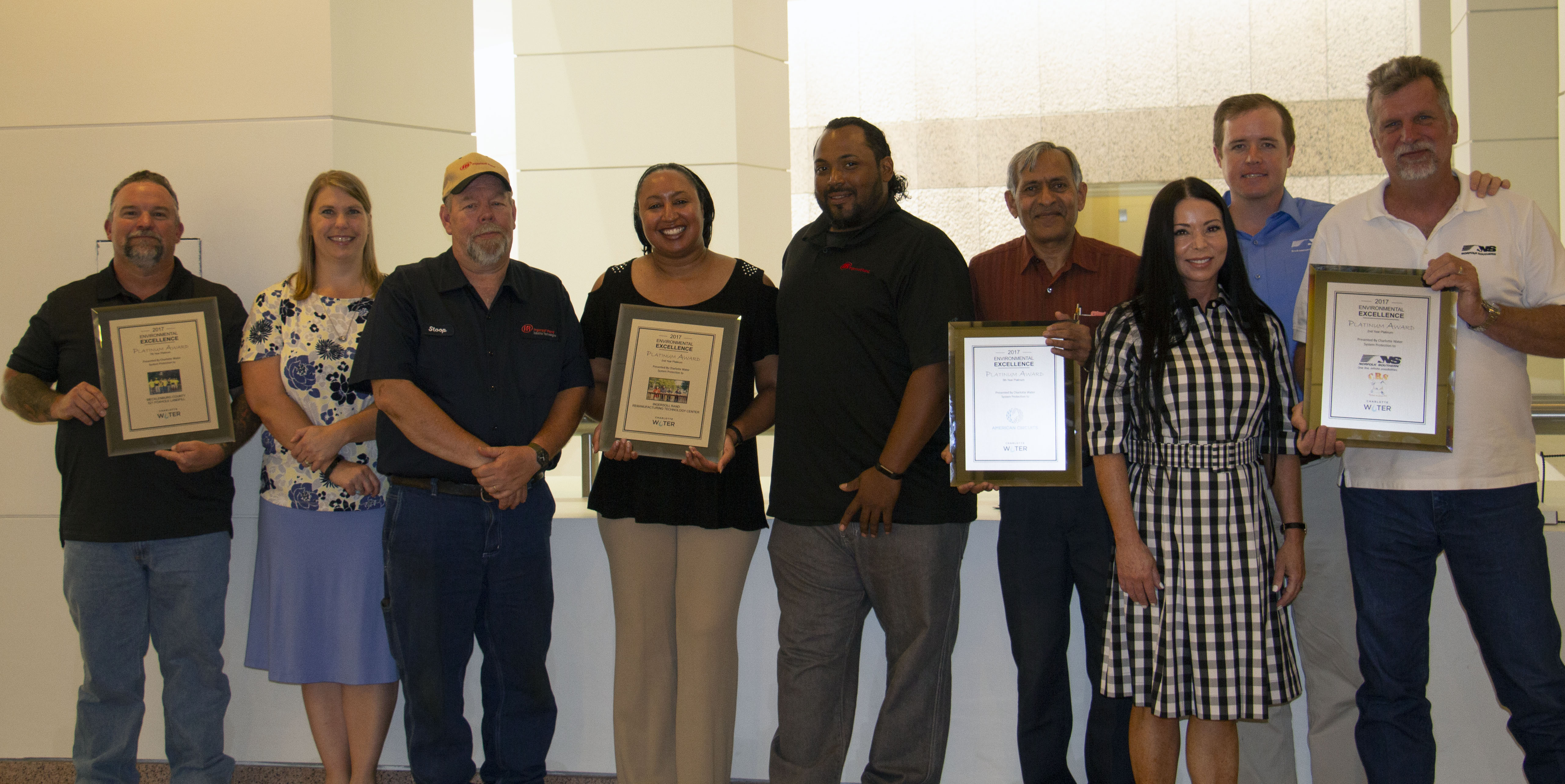 row of 9 utility employees casual office attire stand in a row posing for a group photo in front of an interior stone balcony, 4 of the employees hold framed awards certificates, each with different frame colors