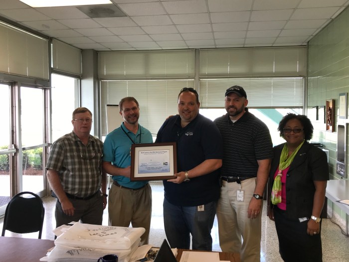 5 utility employees stand in a row in a lobby posing for the camera as the tallest man holds a brown framed certificate