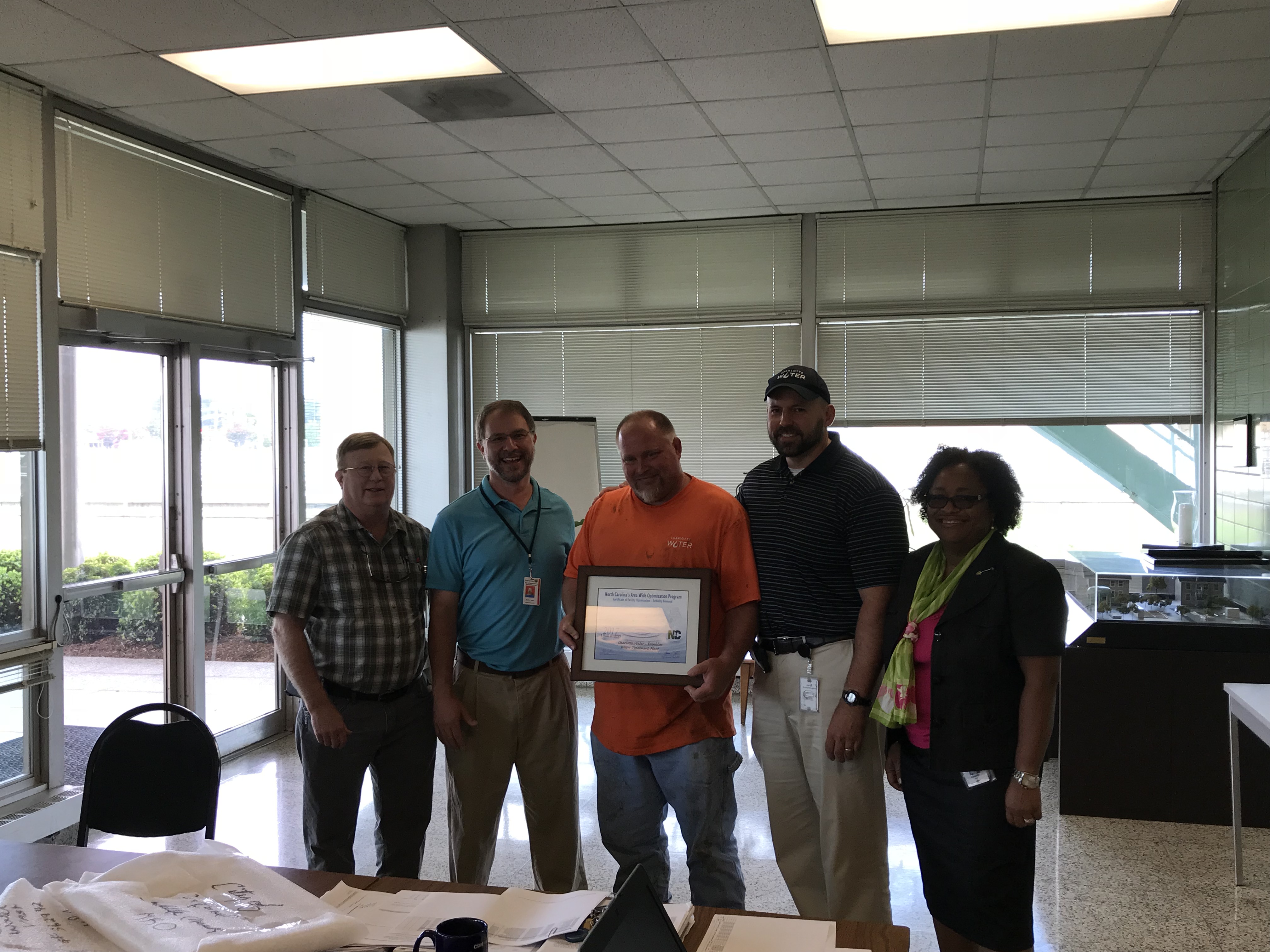 5 utility employees pose for a group photo in a conference room with floor to ceiling windows, 1 employee in a black polo shirt holds a white and tan award in a brown frame