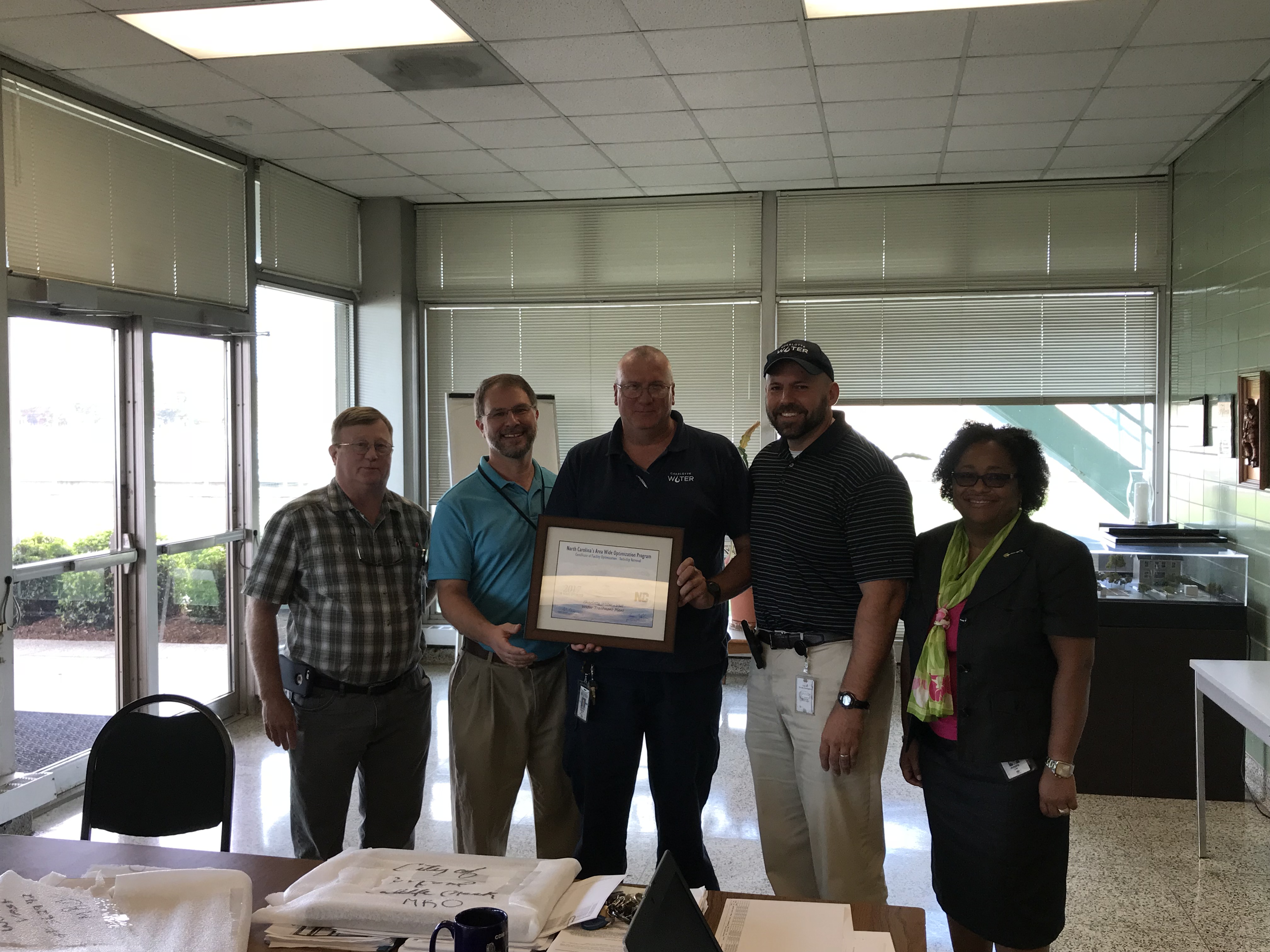5 utility employees pose for a group photo in a conference room with floor to ceiling windows, 1 employee in a black polo shirt holds a white and tan award in a brown frame