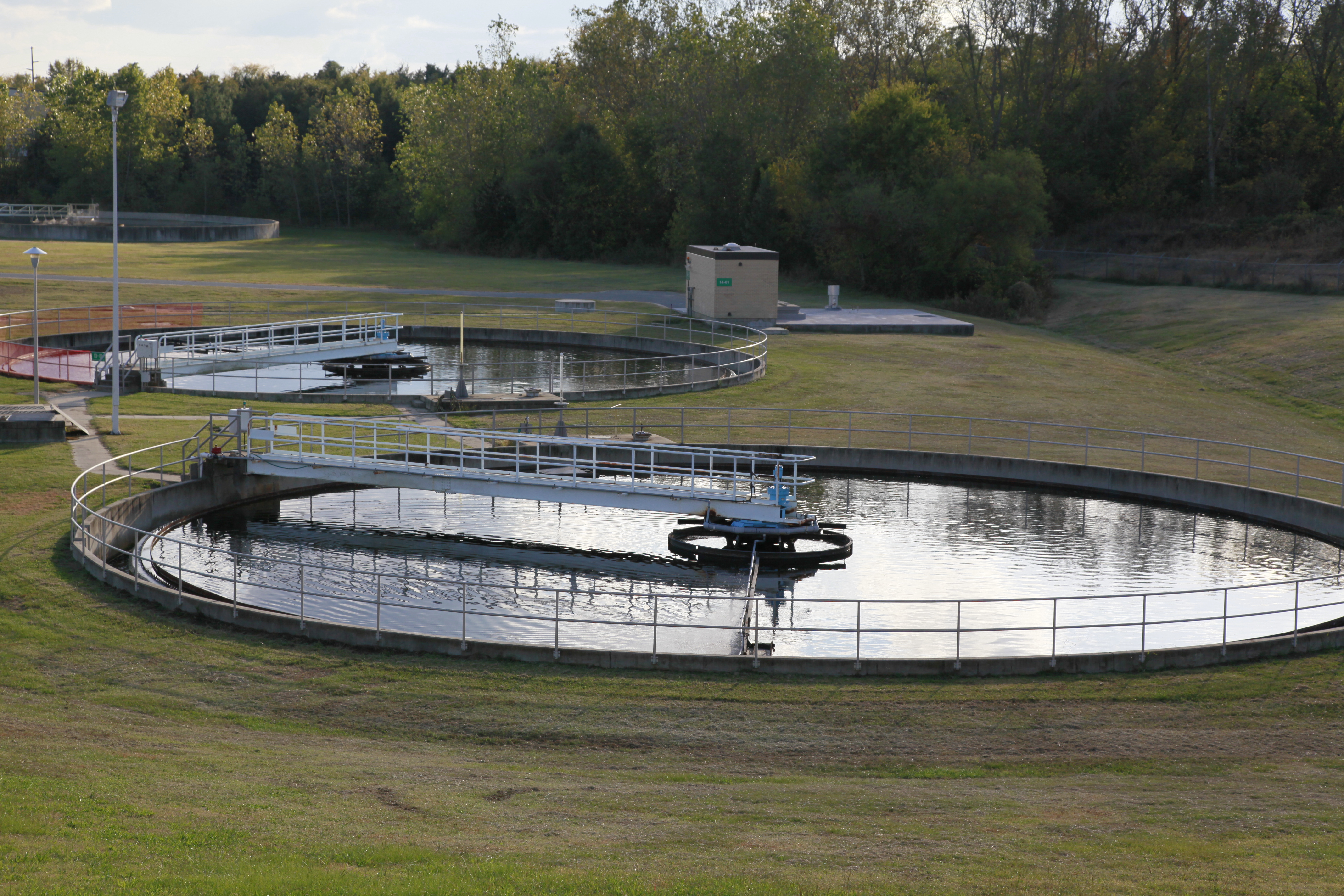birds-eye view of 2 round concreate water treatment pools each with white metal catwalk spanning from center to perimeter; freshly mowed grass spans around them and a small block building on a concrete platform stands in the wooded background