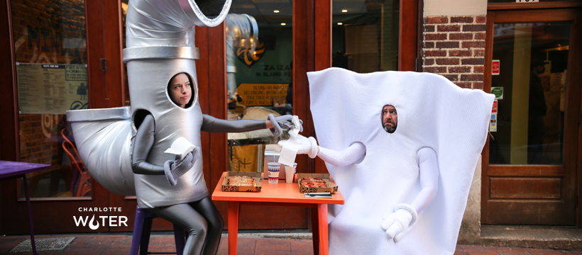 utility mascots woman dressed as a grey metal pipe holding roll of toilet paper vamps at a cafe table with guy dressed as a disposable wipe holding a wipe; the Charlotte Logo appearing in white lettering towards bottom of image