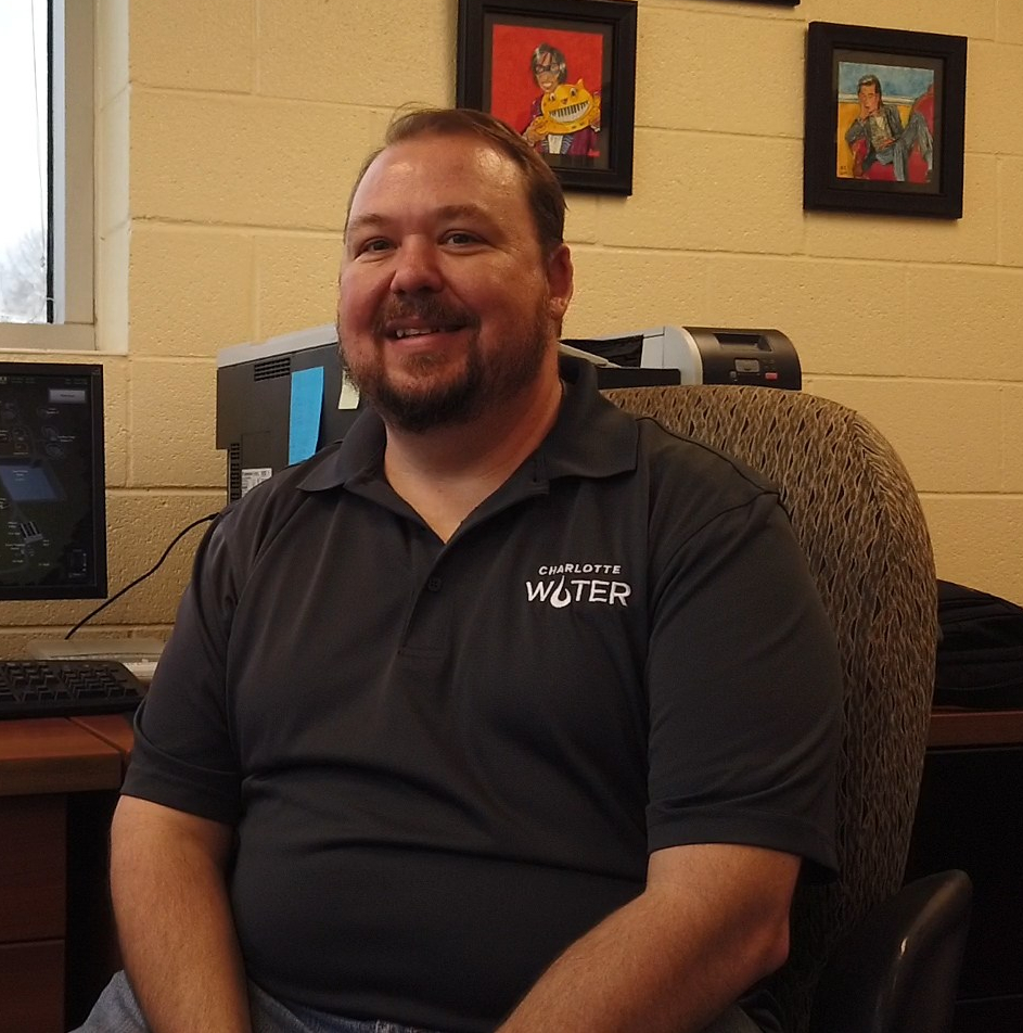 utility employee in black polo shirt with Charlotte Water logo sits in a chair at a desk in an office