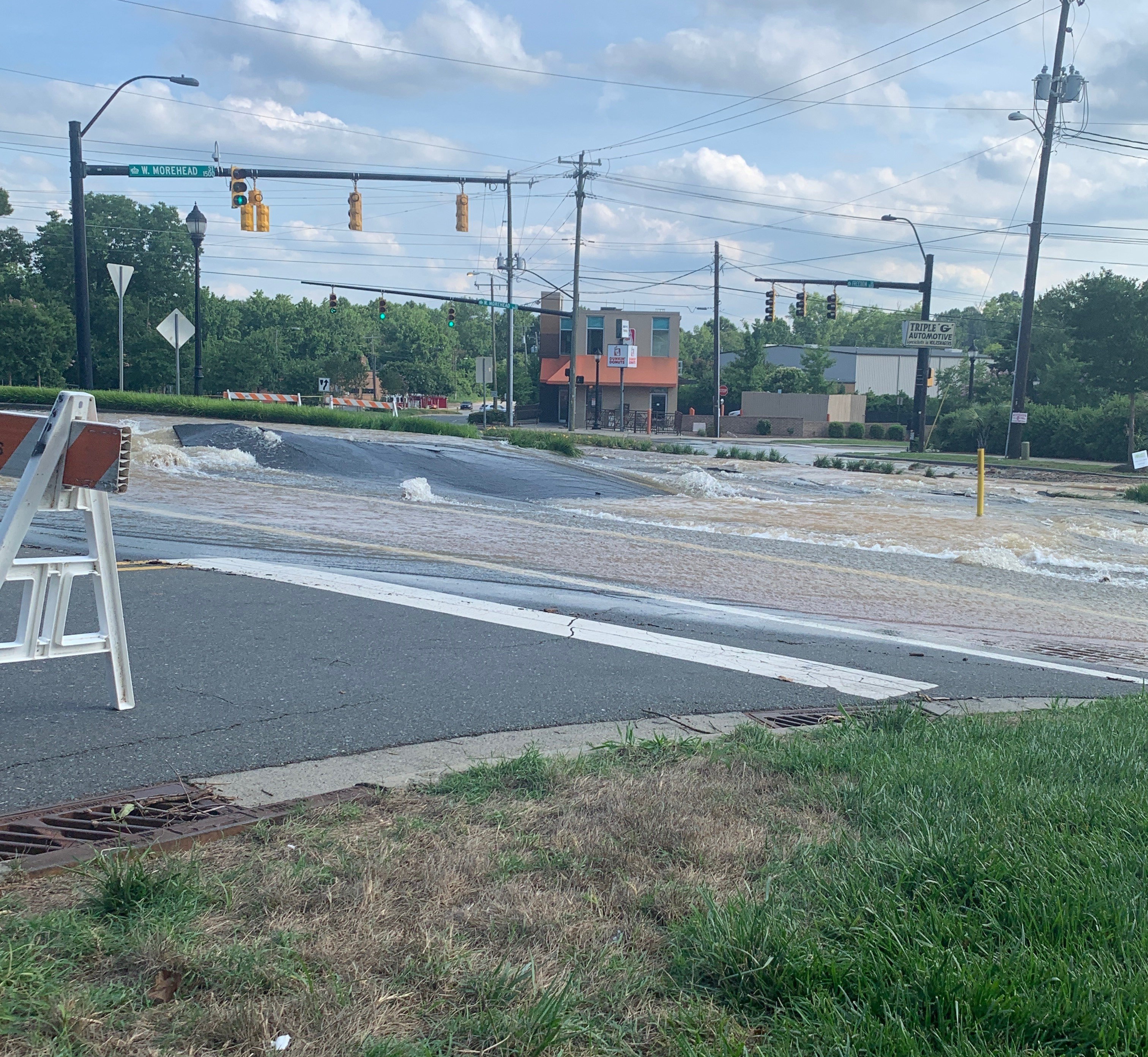 muddy waters roar down urban street flooding an intersection; 4 orange and white saw horses are stationed nearyby