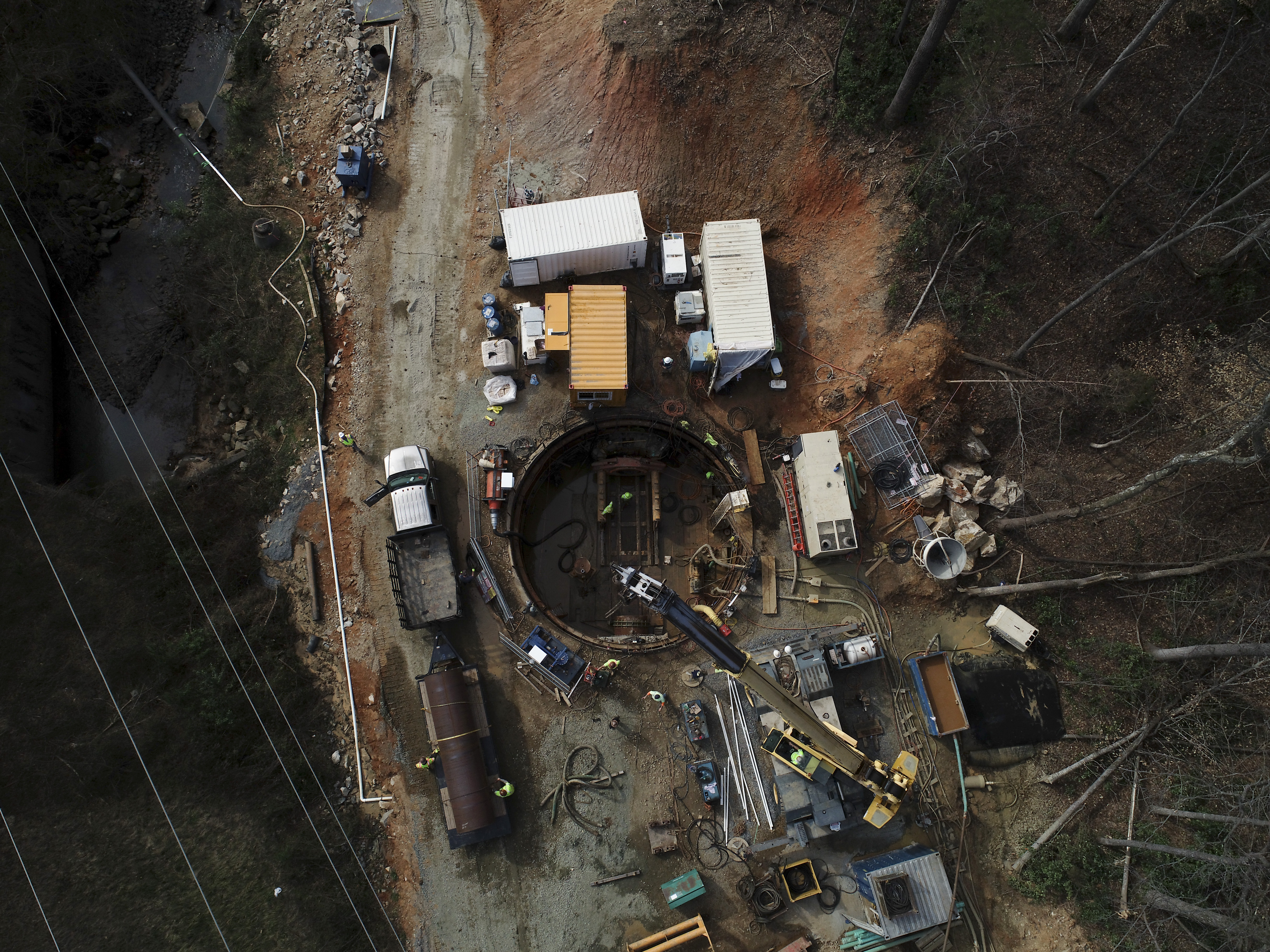 birds-eye-view of recently excavated construction site with 3 cargo containers, 1 generator truck, yellow excavator, 1 white stake bed truck, water tanker trailer; everything surrounds an old steel lined, giant round cistern in the ground. Nearby is a creek with eroded dirt embankment and other construction gear
