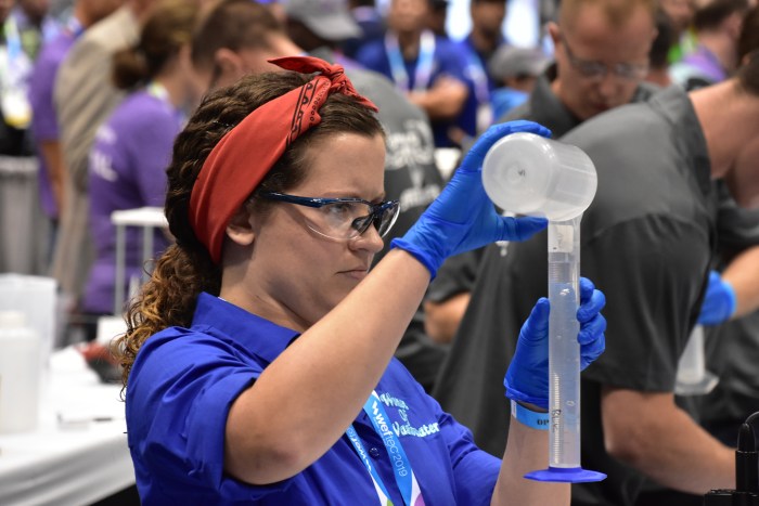 utility employee wearing blue shirt and red bandana headband demonstrates at a crowded tradeshow how to pour water from a sample jar into a tall, cylindrical measured plastic beaker