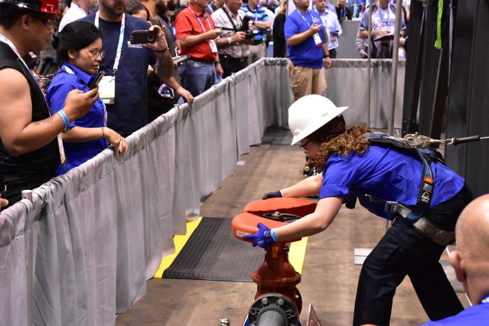 at a tradeshow, a utility department technician in a blue shirt, white hard hat, blue glove and a black safety harness, demonstrates to the crowd of attendees standing behind a grey draped divider, how install a red safety cover onto a the wheel of pipe valve