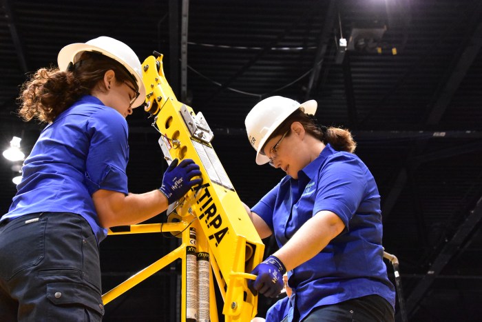 2 utility employees wearing blue shirts, white hard hats, pony tails and safety glasses demonstrate at a tradeshow the operation of a yellow XTIRPA ballast arm for the utility technician out in the field