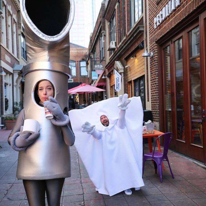 2 utility company mascots vamp in front of a row of shops: 1 dressed up as a piece of metal pipe slurps beverage with a straw holding a roll of toilet paper while the second mascot is dressed a a white sheet of disposable wipes